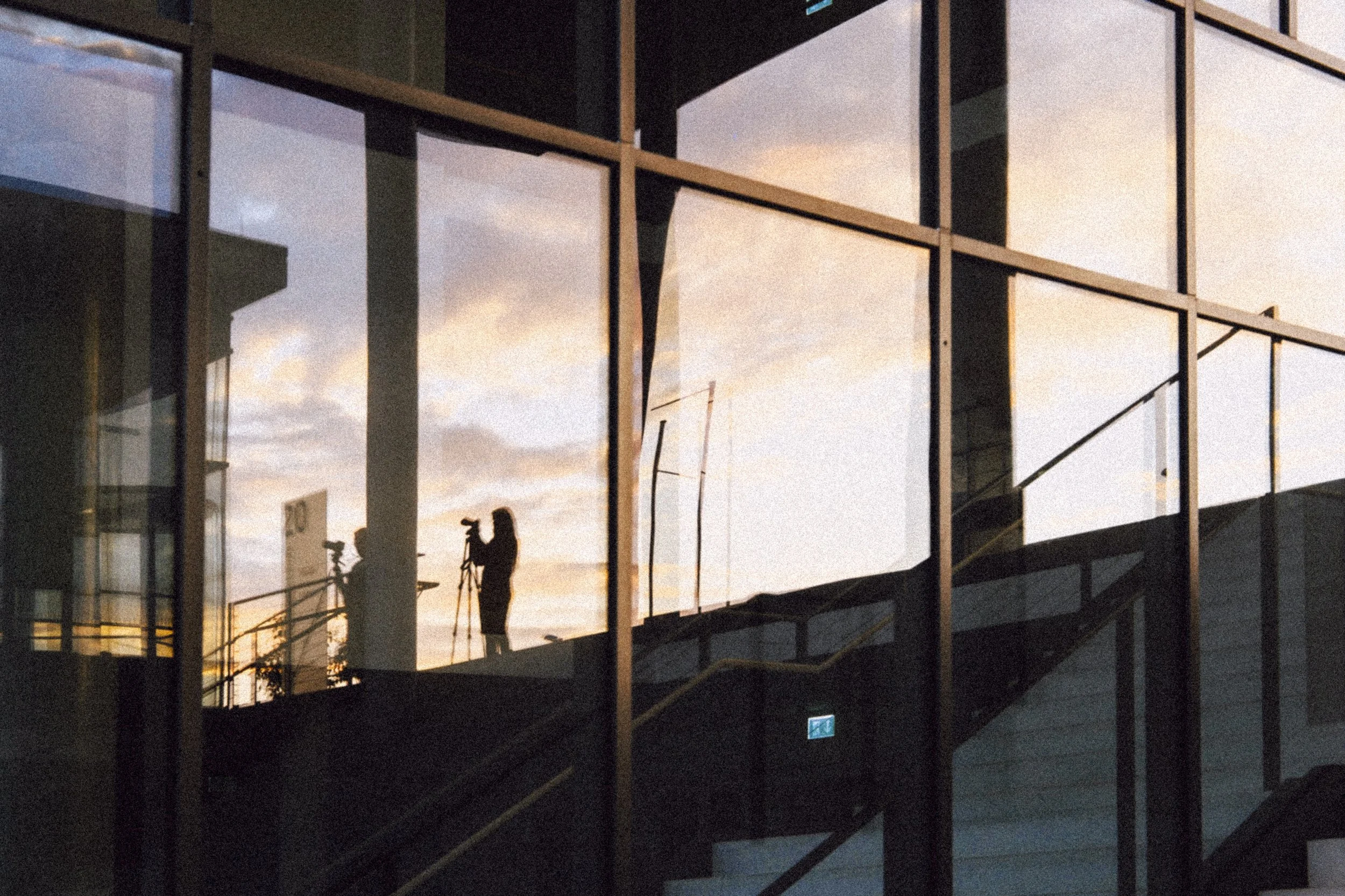 Silhouette of a person setting up a camera on a tripod inside a building with large reflective glass windows, with a sunset sky in the background.