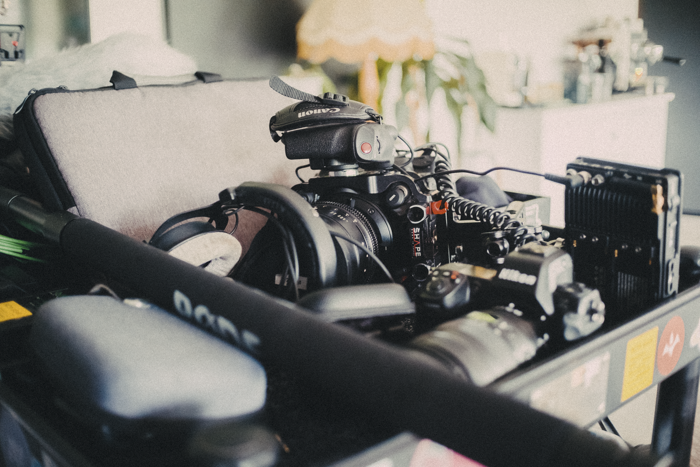 Close-up of a professional camera mounted on a rig with headphones, a microphone, and various accessories, on a black cart in a room with a beige lamp and green plant in the background.
