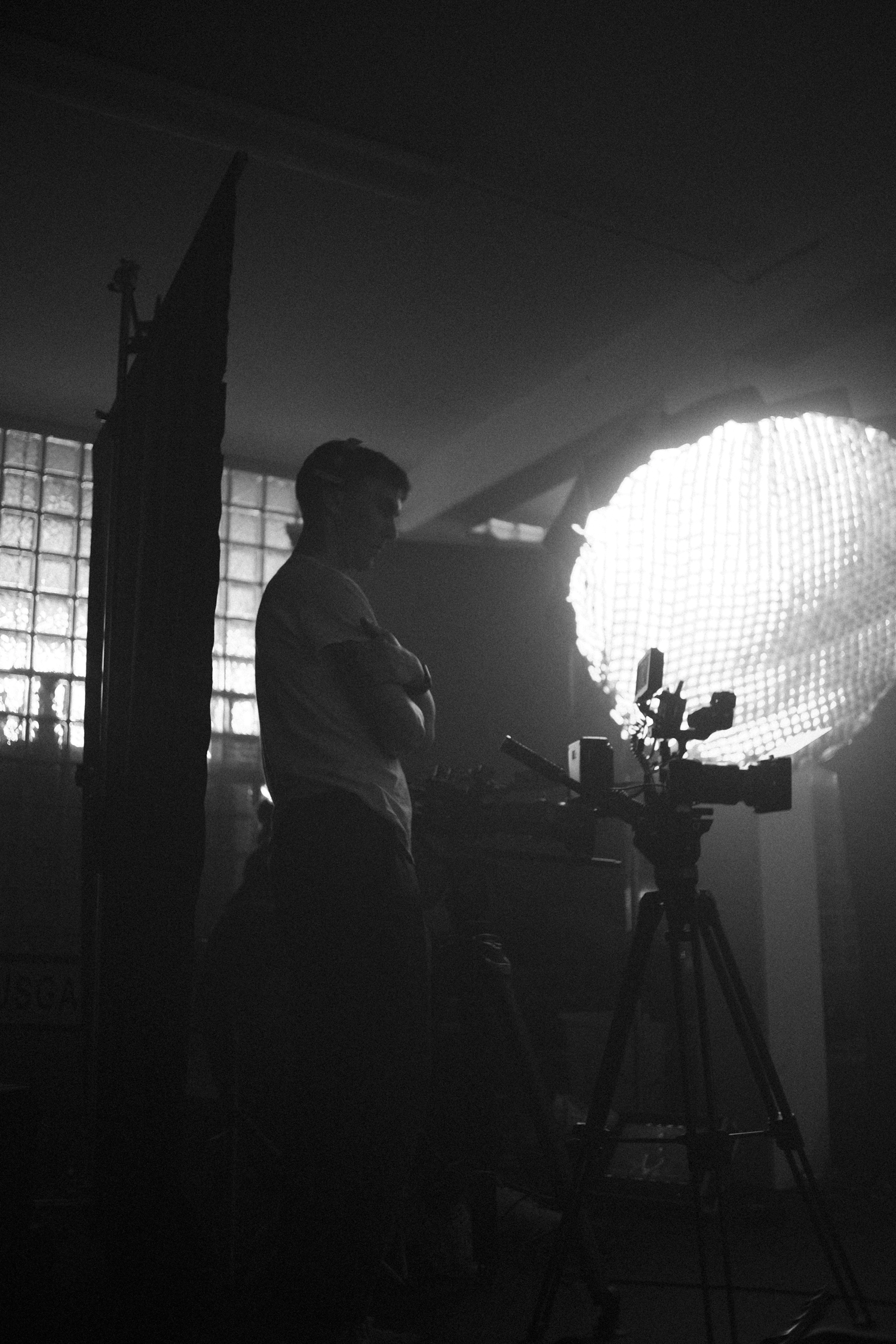 Silhouette of a person standing next to a camera on a tripod, with bright lights and windows in the background.
