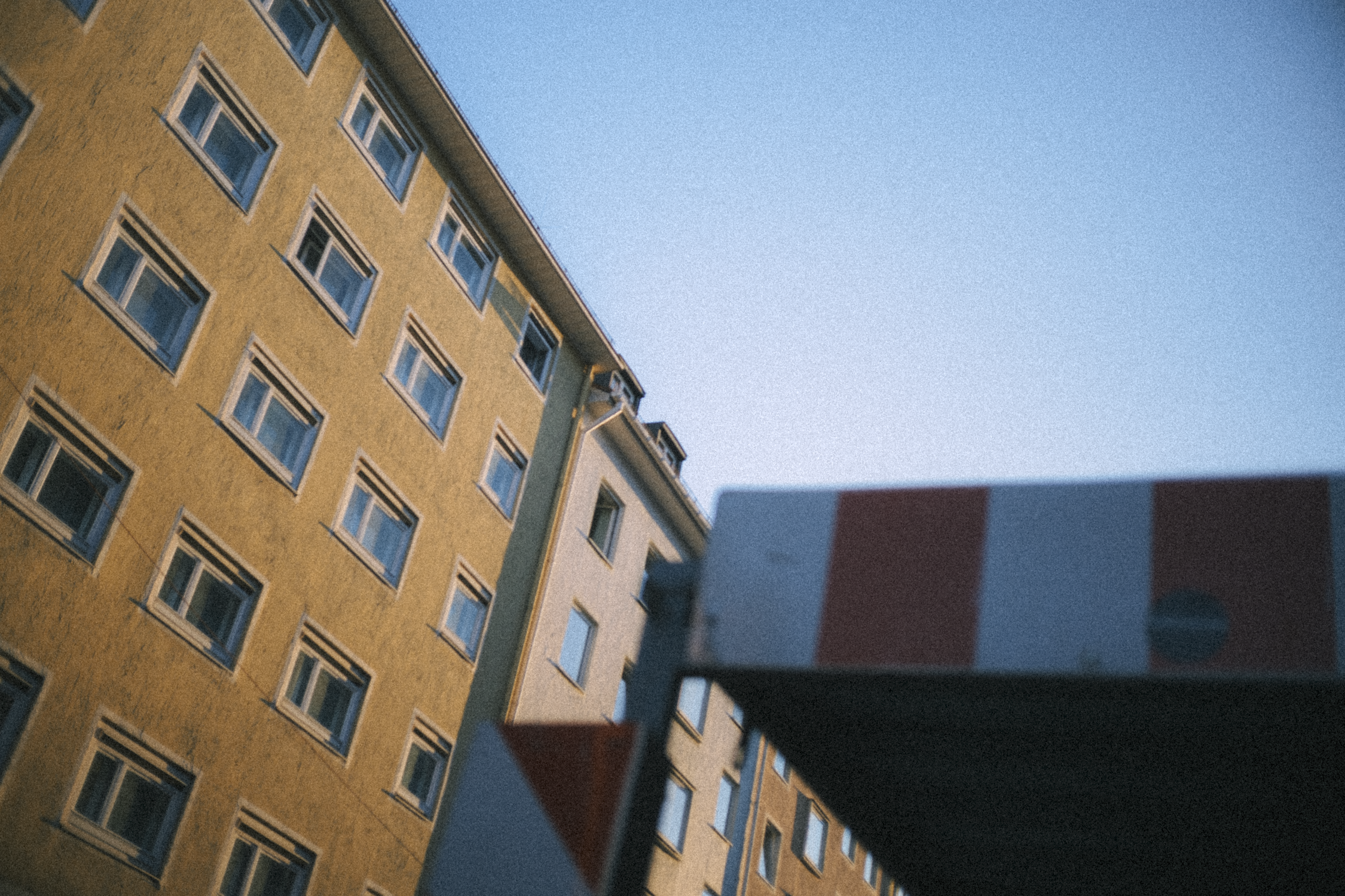 Low-angle view of a yellow multi-story residential building with multiple windows, against a clear blue sky. Part of a red, white, and blue sign is visible in the foreground.