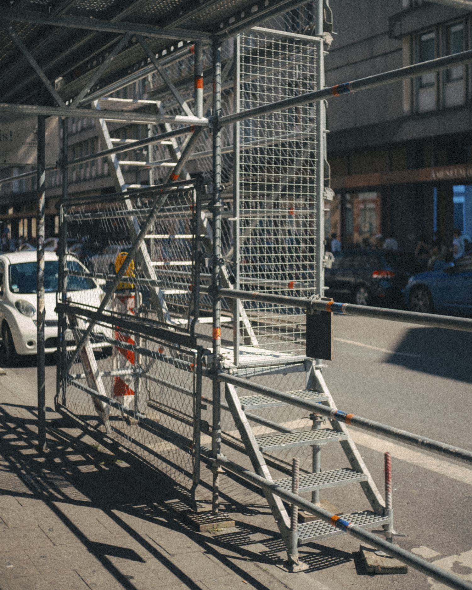 Scaffolding and stairs on a city sidewalk with parked cars and buildings in the background.