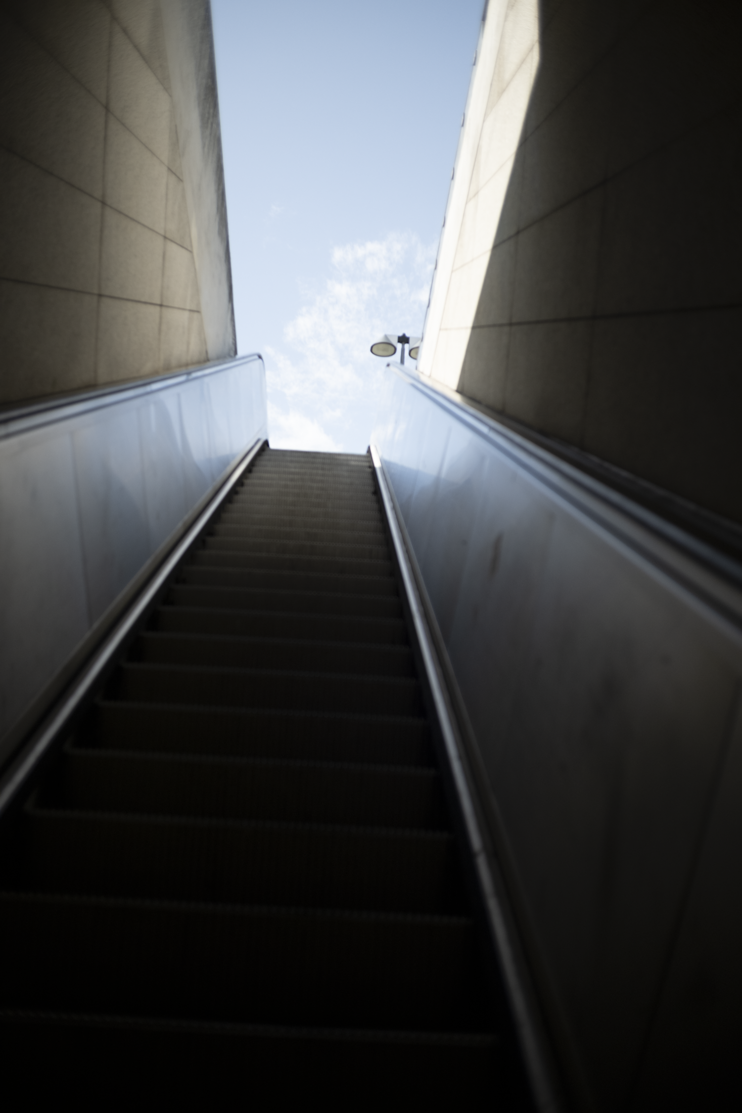 Downward view of an escalator leading upwards towards a bright blue sky with a few clouds, framed by concrete walls.