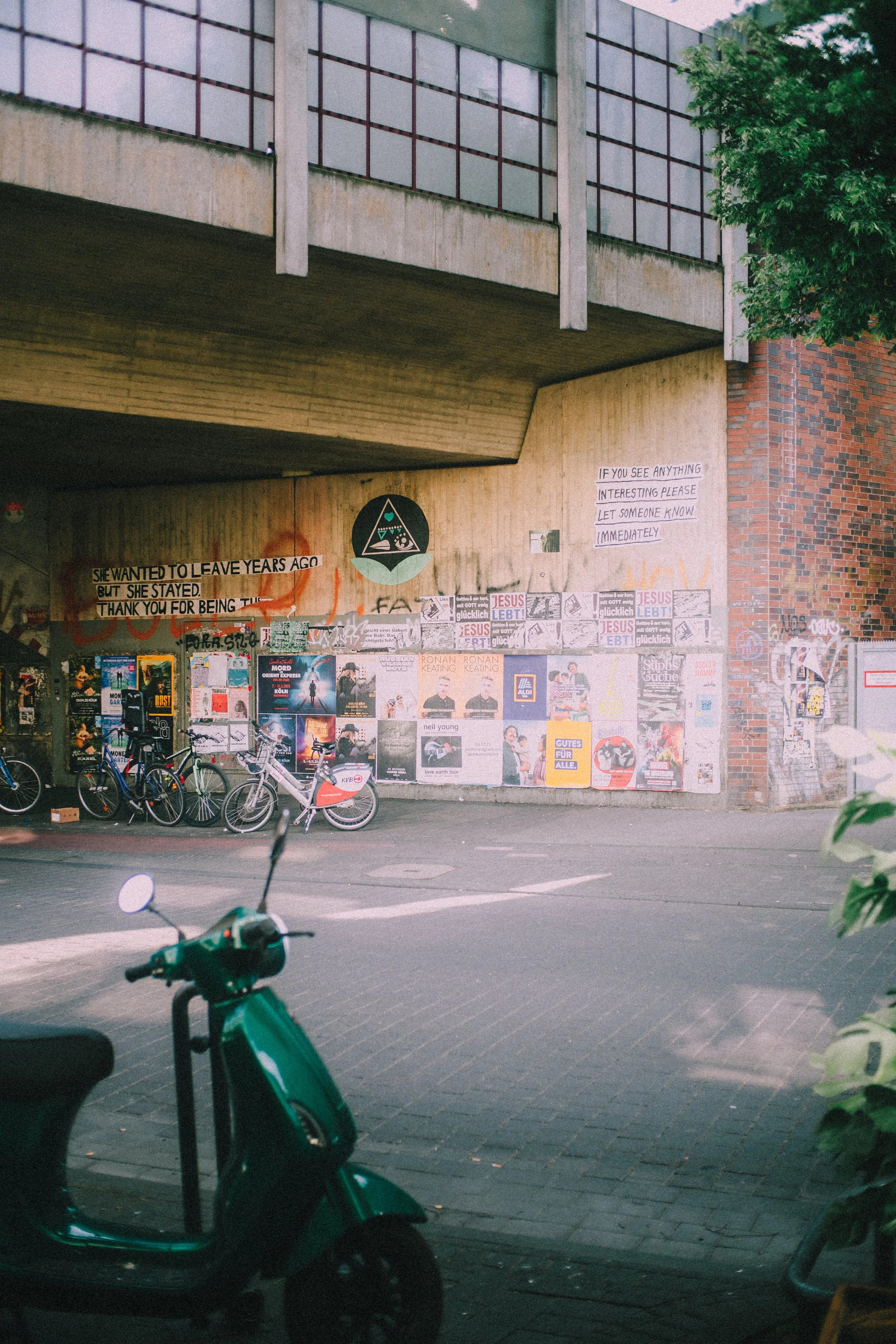 Graffiti-covered wall under a bridge with posters, stickers, and signs, with bicycles and a scooter parked in front, and a tree on the right side.