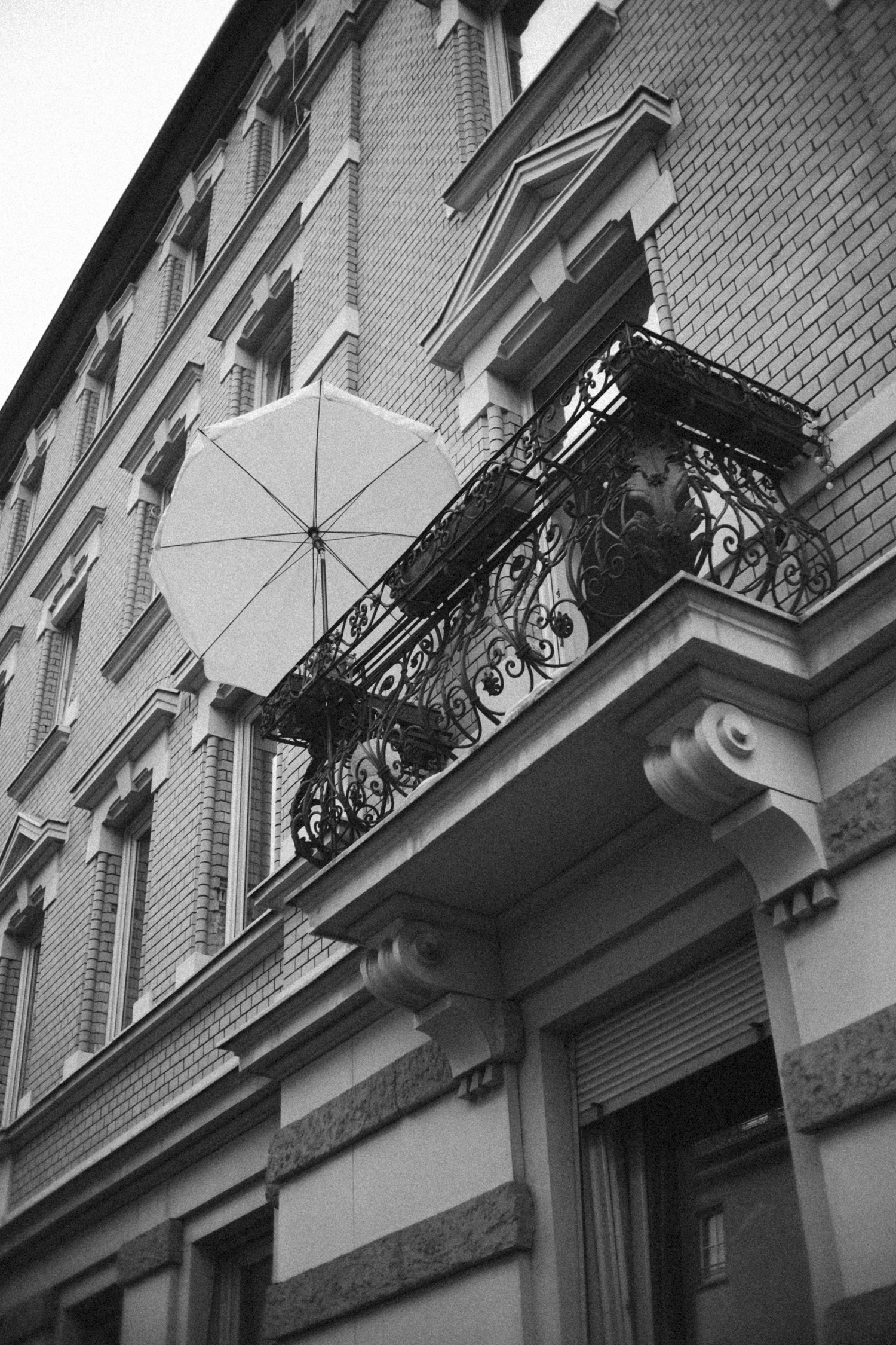 Black and white photo of a building with a small ornate balcony that has a wrought iron railing. A closed umbrella is placed on the balcony. The building has decorative window trim and brickwork.