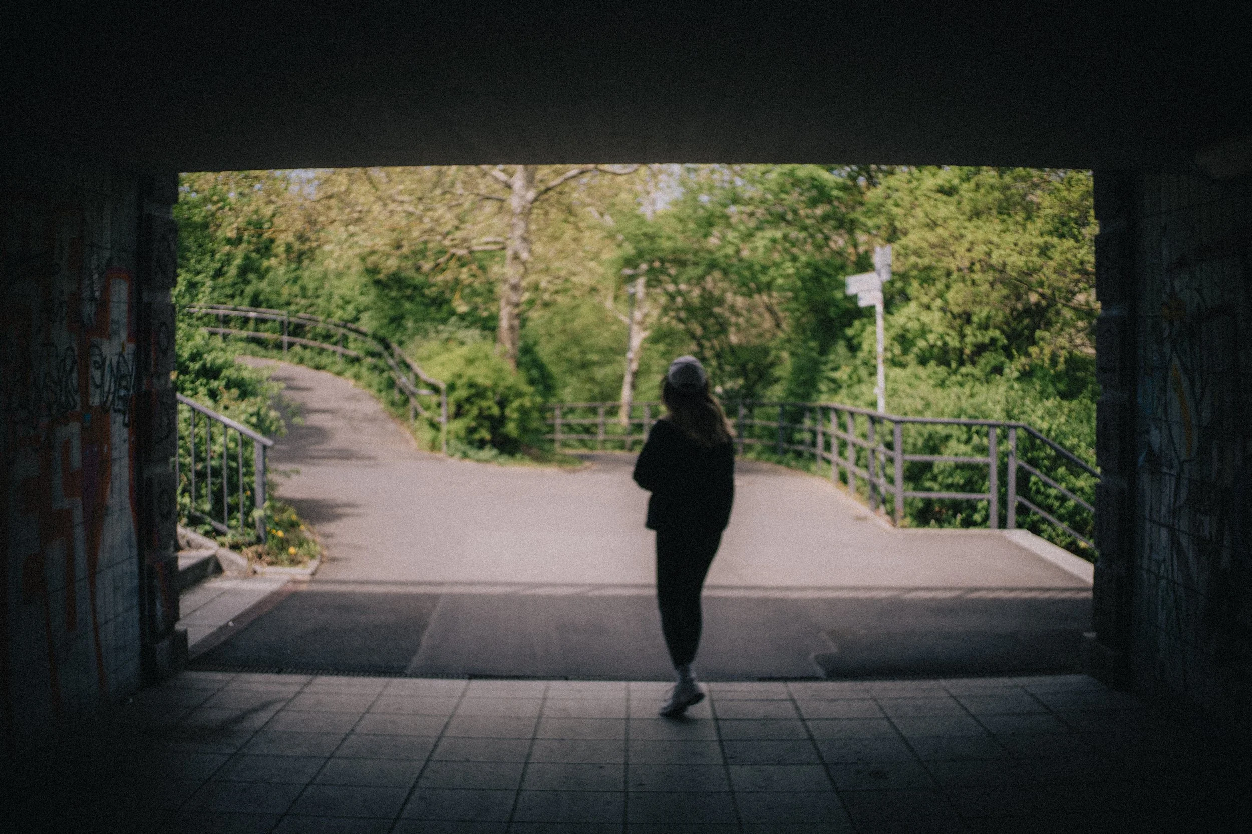 Silhouette of a person walking out of a tunnel underpass into a green, wooded park area.