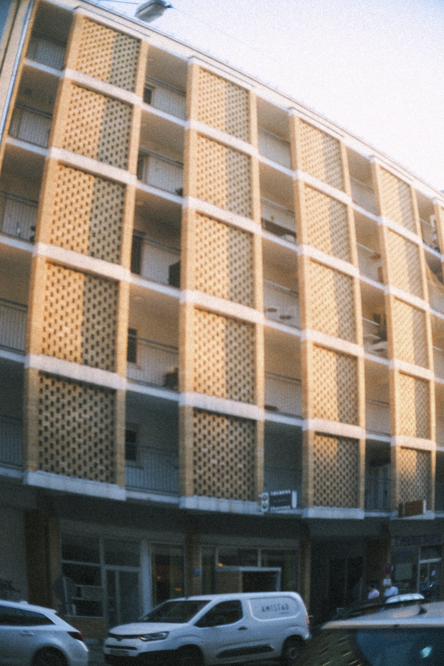 Multi-story building with a patterned facade, parked cars in front, and a few people near the entrance.