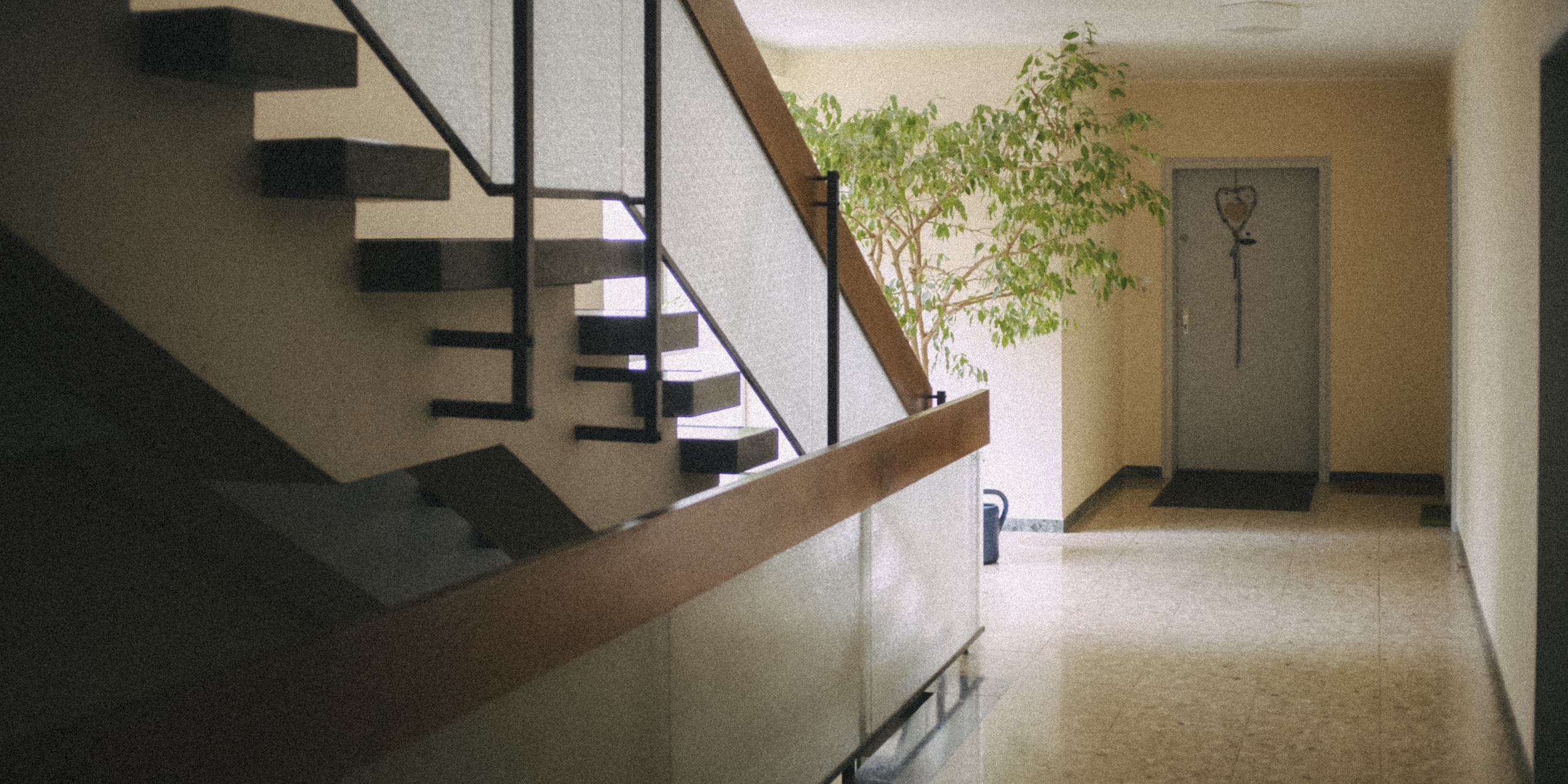 Interior of a building with a staircase on the left, a potted plant at the top of the stairs, and a doorway with a heart-shaped decoration on the door in the back. The floor is made of speckled tiles.