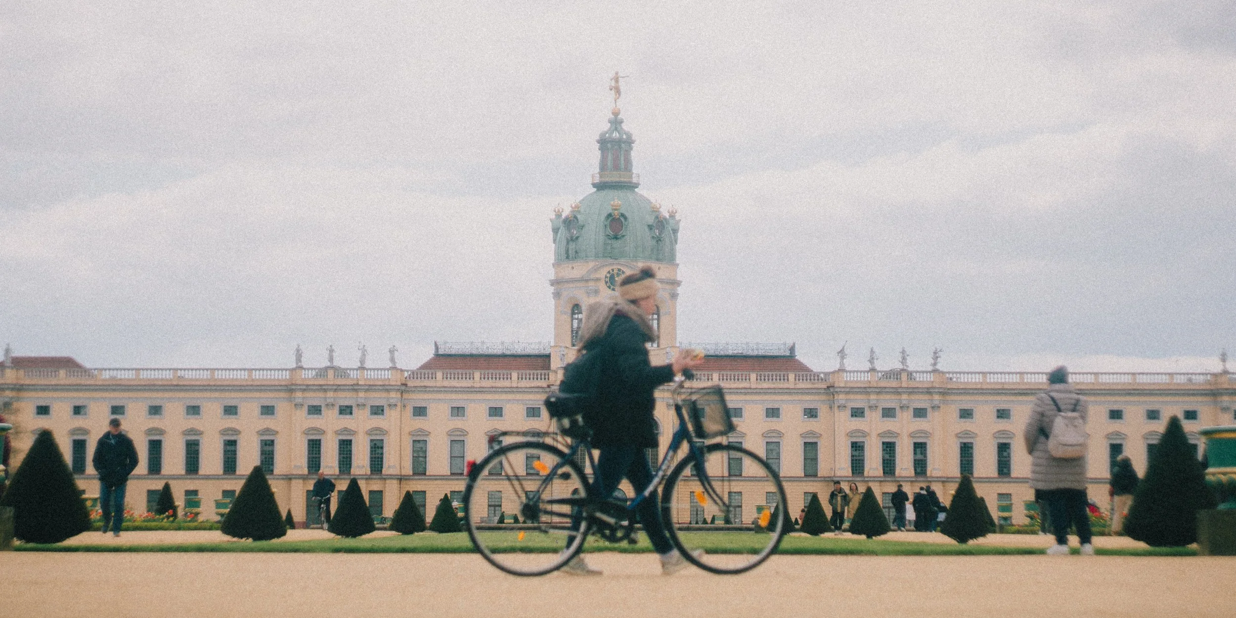 A person riding a bicycle in front of a historical building with a clock tower, surrounded by people and manicured gardens.