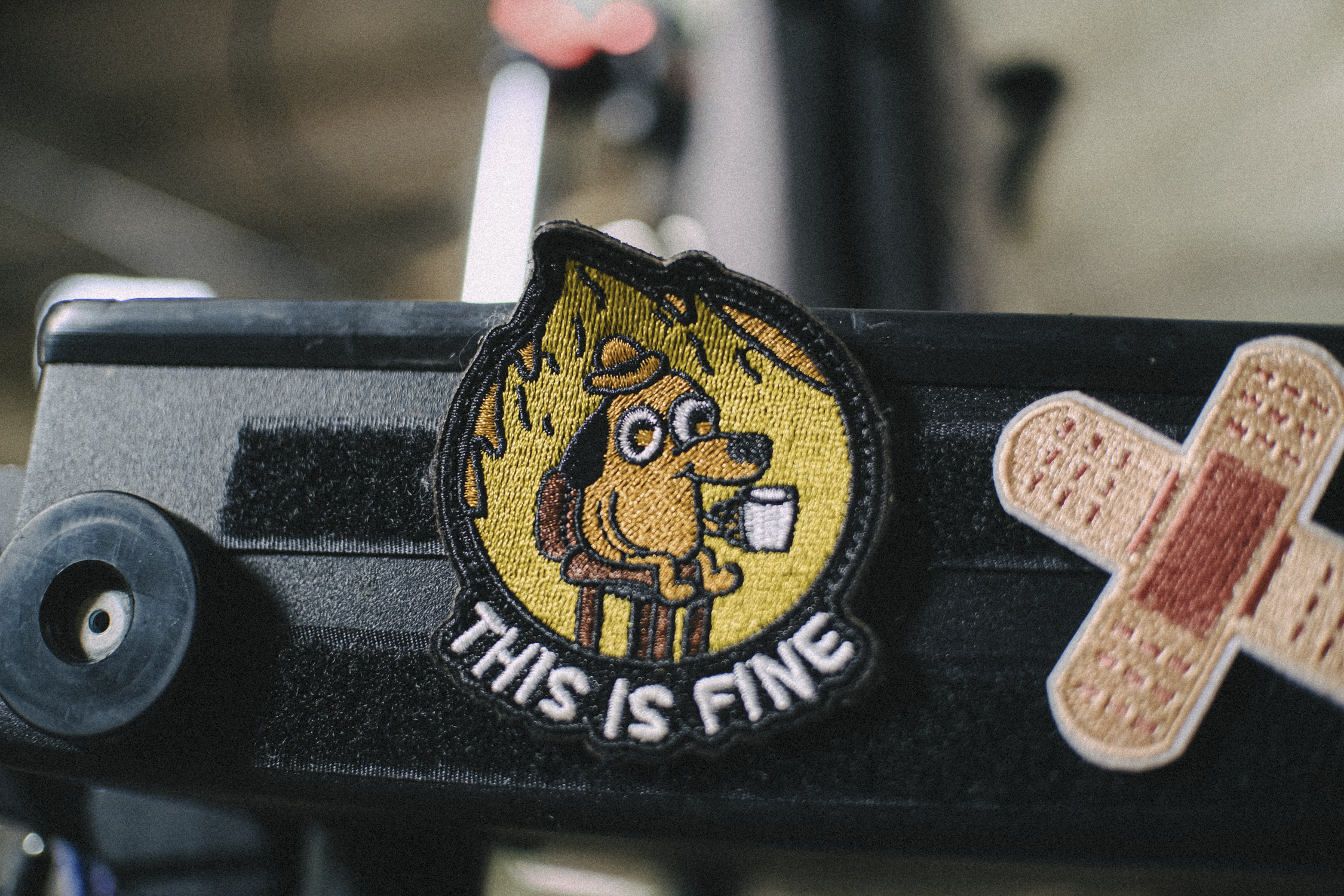 A close-up of two embroidered patches on a dark surface. The central patch features a cartoon dog with a crown, sitting on a chair, holding a mug, with the phrase 'THIS IS FINE' below. The second patch, partially visible, resembles a bandage.