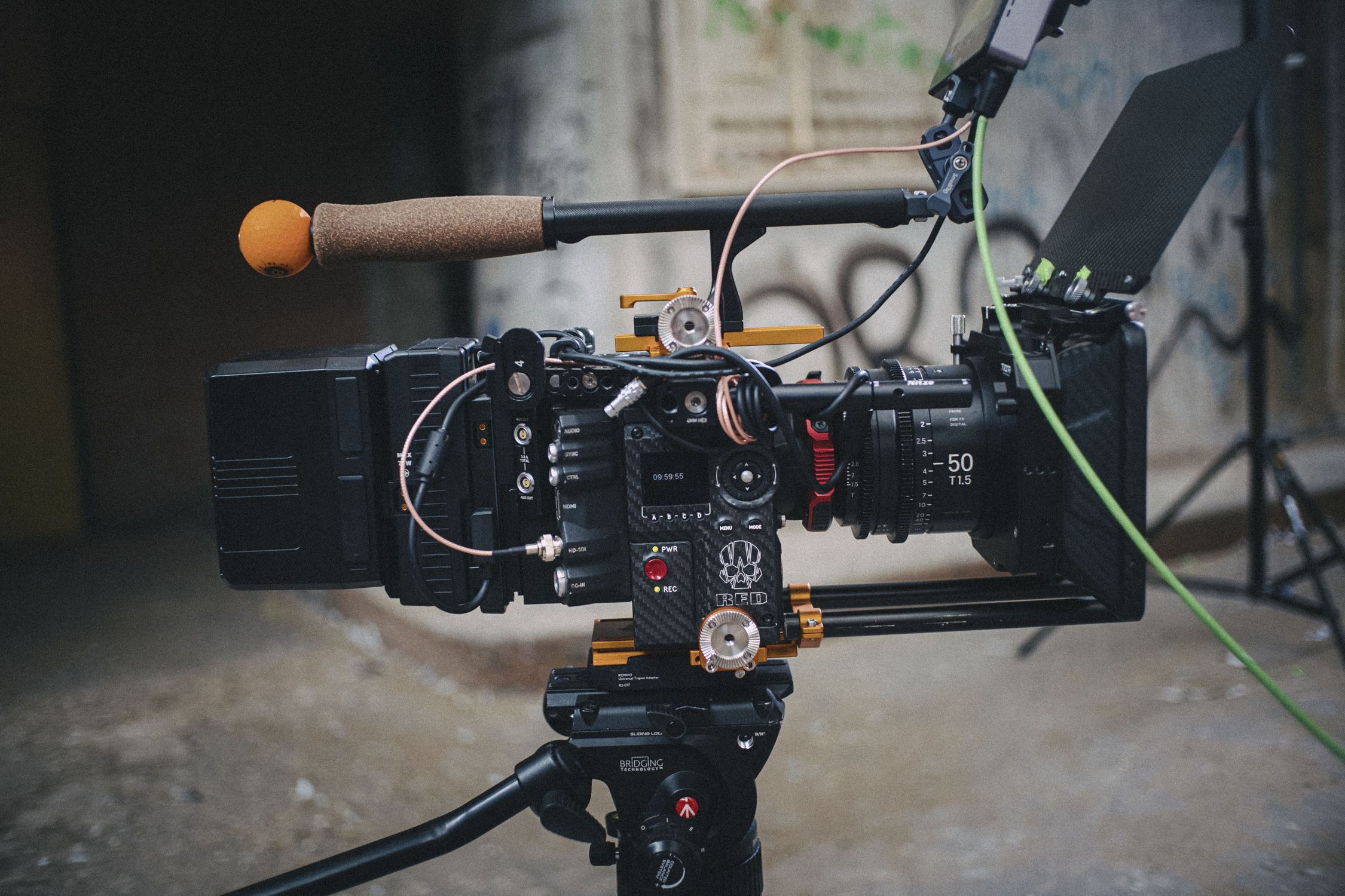 A professional video camera mounted on a tripod with various attachments, including a shotgun microphone, wires, and controllers, positioned in a space with graffiti on the wall.