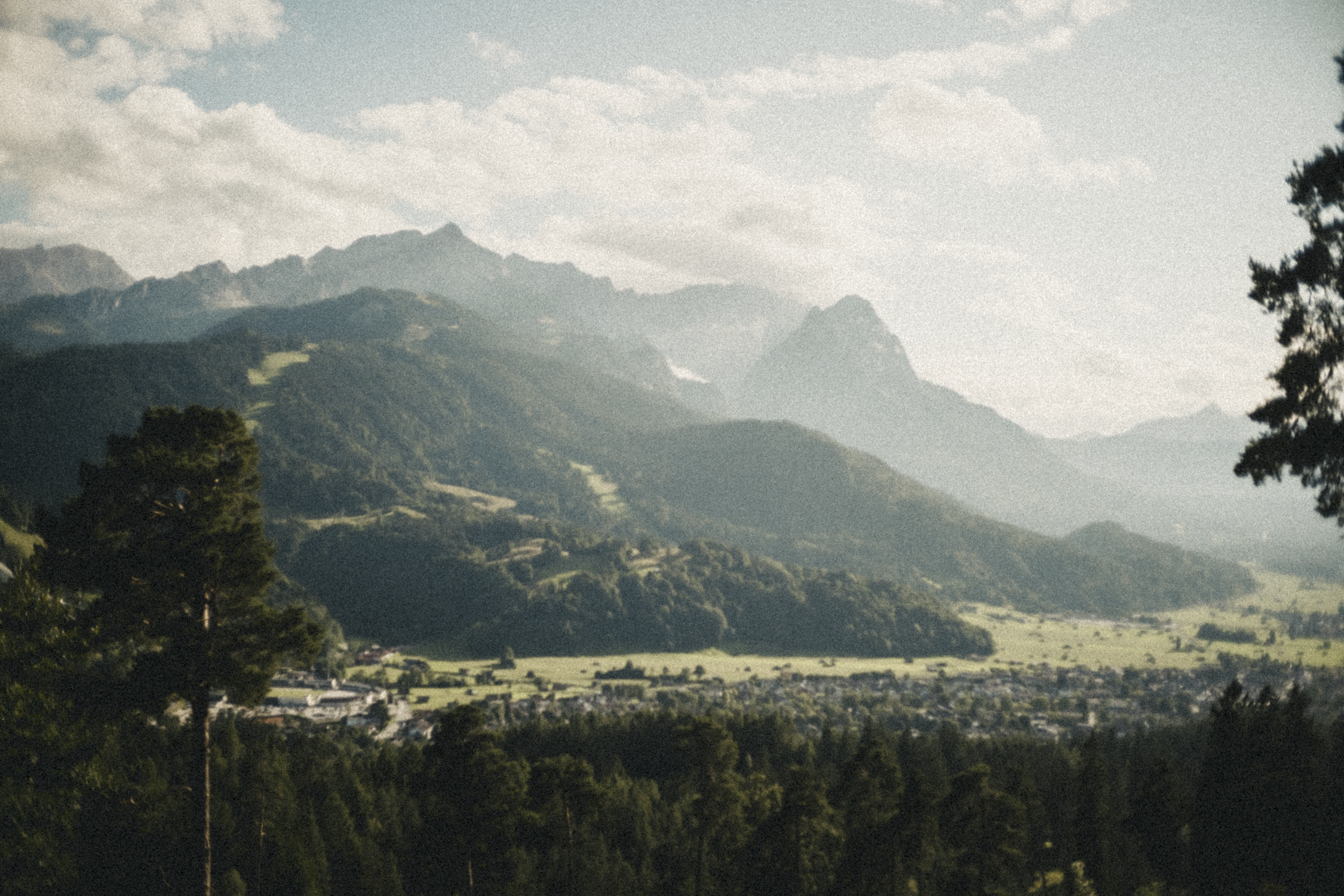 A scenic view of a mountain range with lush green forests and a small town in the foreground under a partly cloudy sky.
