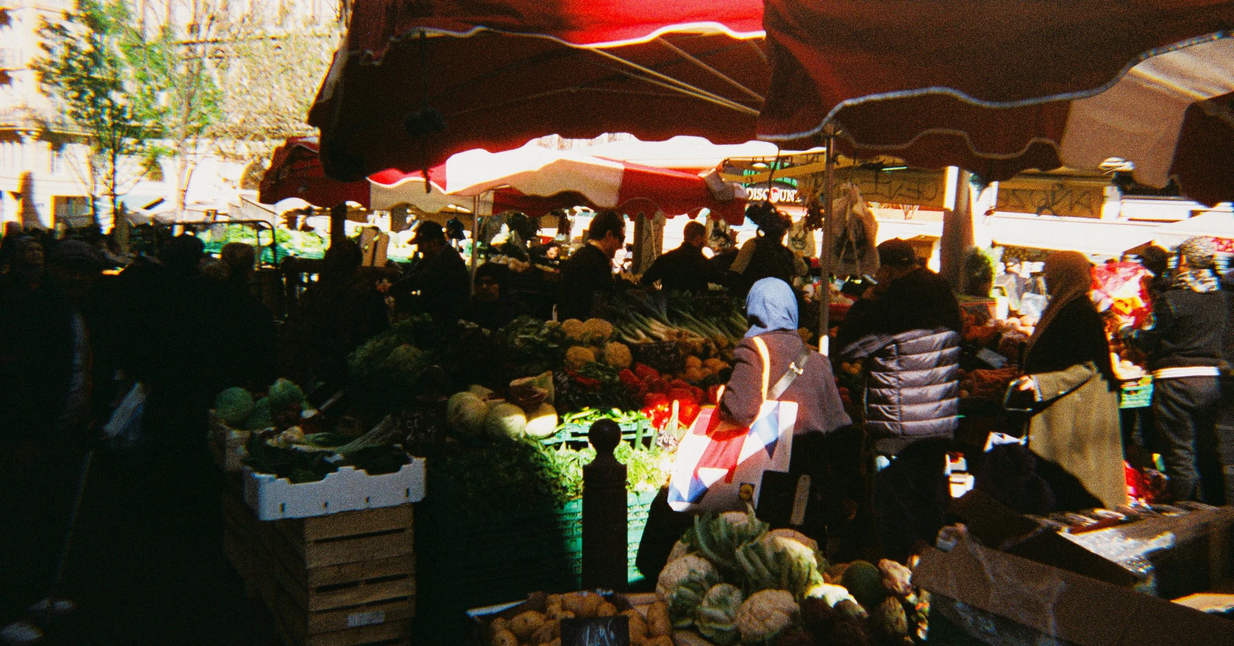 Outdoor farmers market with vendors selling vegetables and shoppers browsing under red and white striped tents.