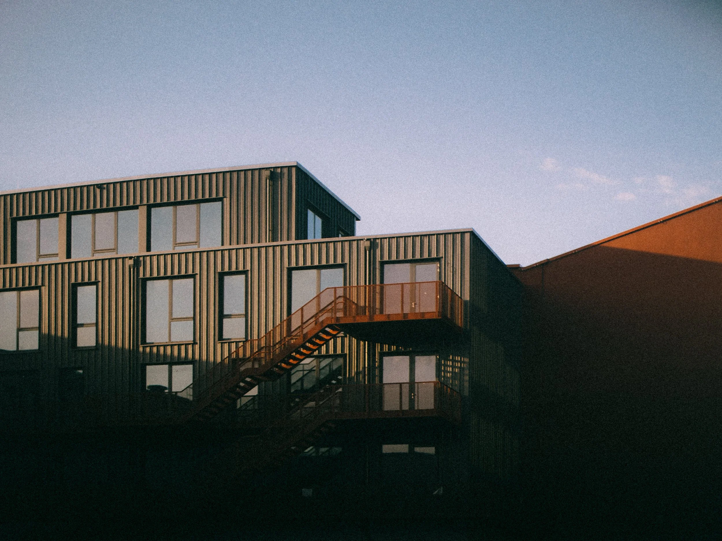 Modern multi-story building with green and red exterior, metal siding, and orange fire escape stairs against a clear sky.