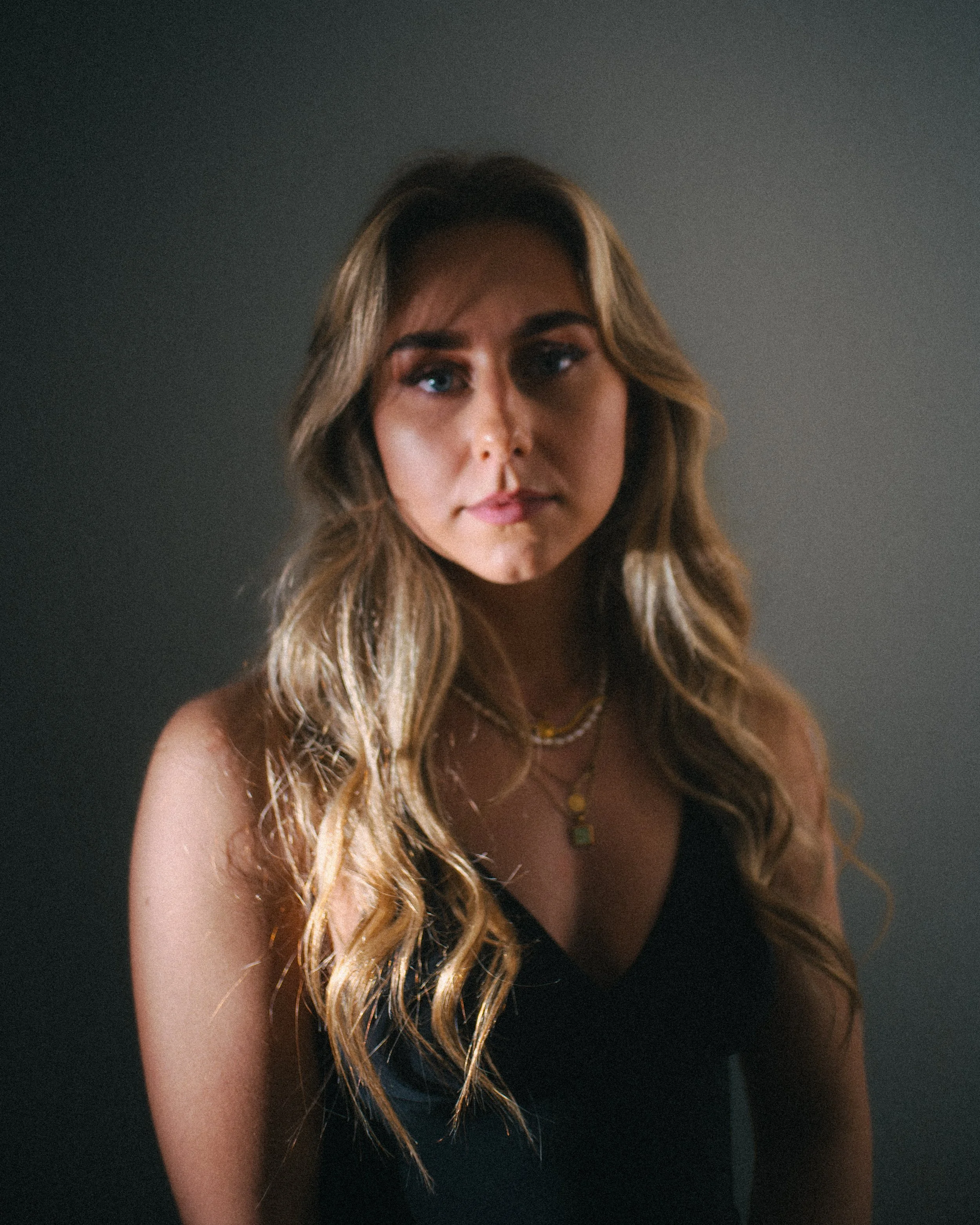 A young woman with long wavy blonde hair wearing a black top and layered necklaces, looking into the camera against a plain gray background.