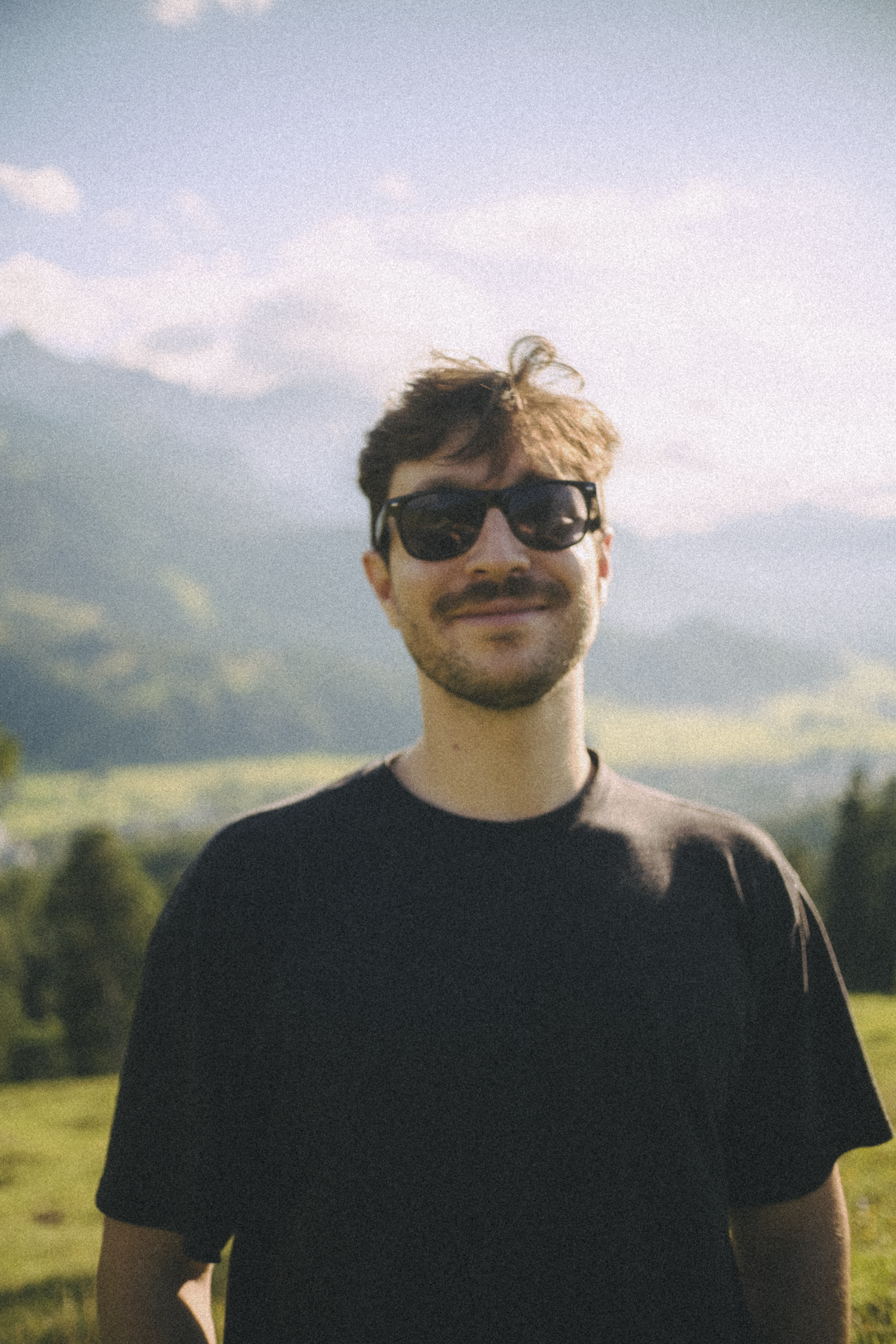 A young man with dark hair and a beard wearing black sunglasses and a black t-shirt outdoors with a mountain landscape and blue sky in the background.