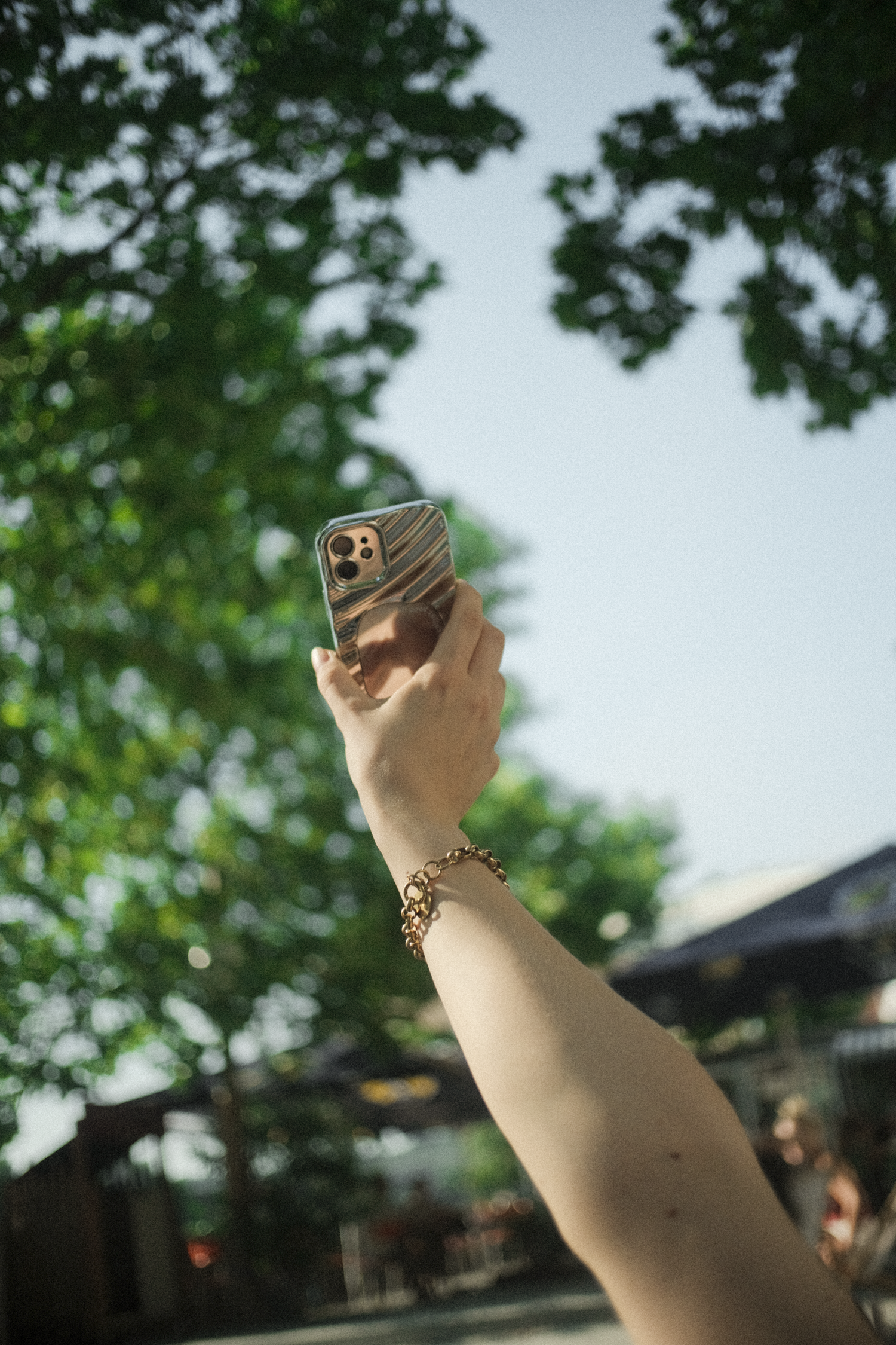 A person's arm holding up a smartphone with a reflective, colorful case outdoors, with green trees and a building in the background.
