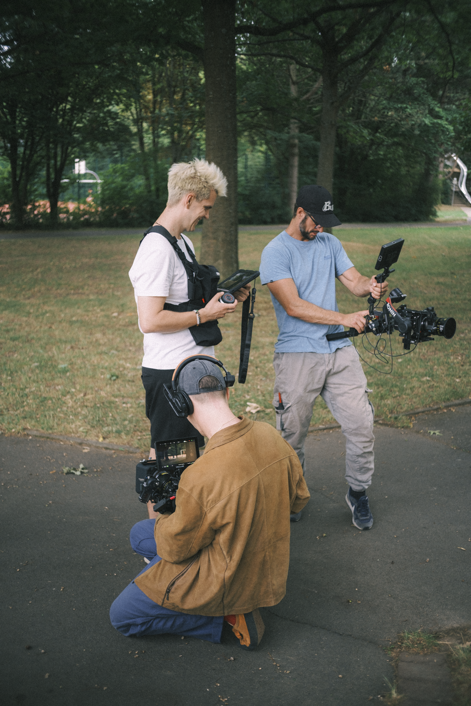 Three people filming in a park, two standing and one kneeling, with trees in the background.