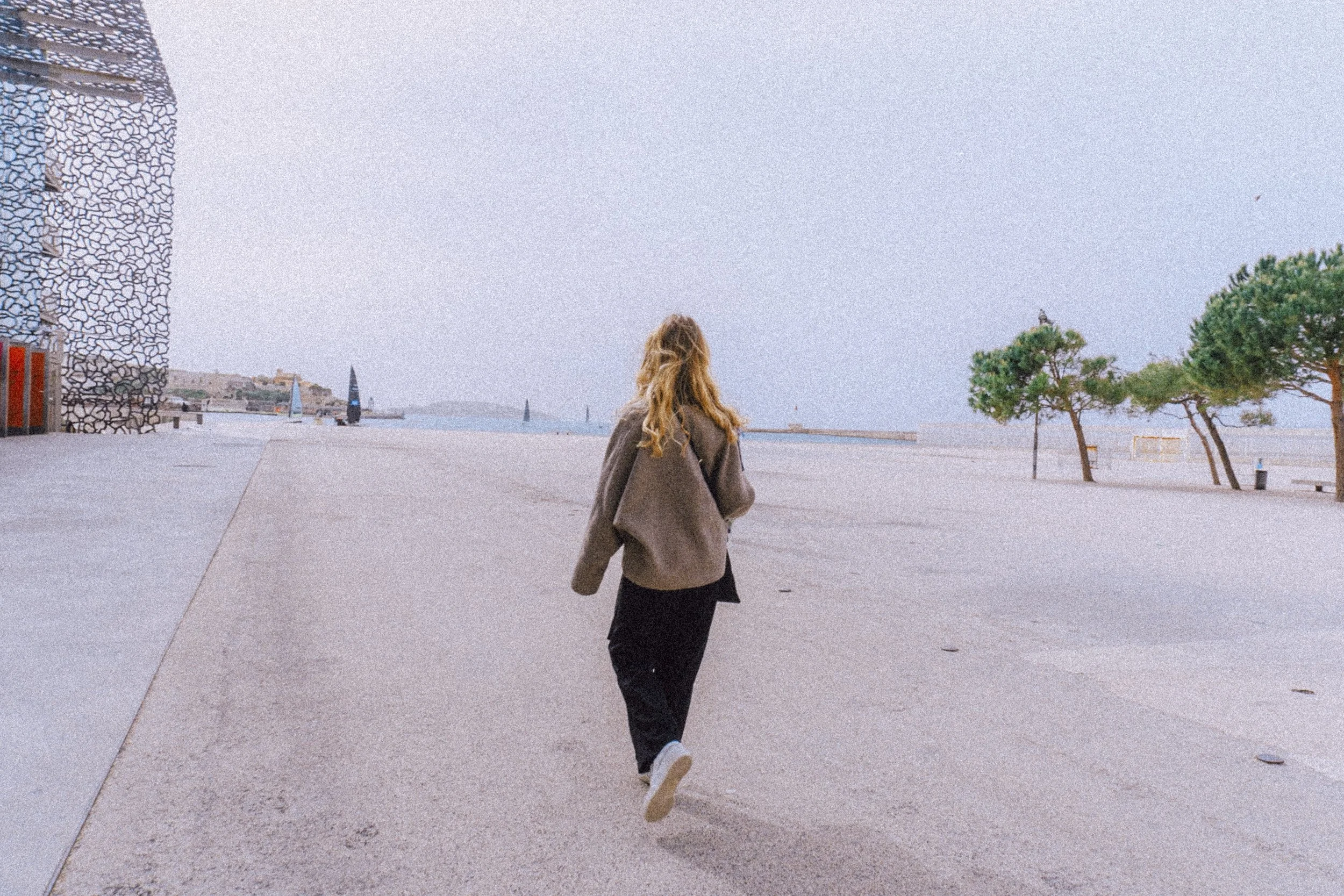 A person walking alone on a wide, empty concrete promenade near the water, with a few trees and modern buildings in the distance, under a light blue, partly cloudy sky.