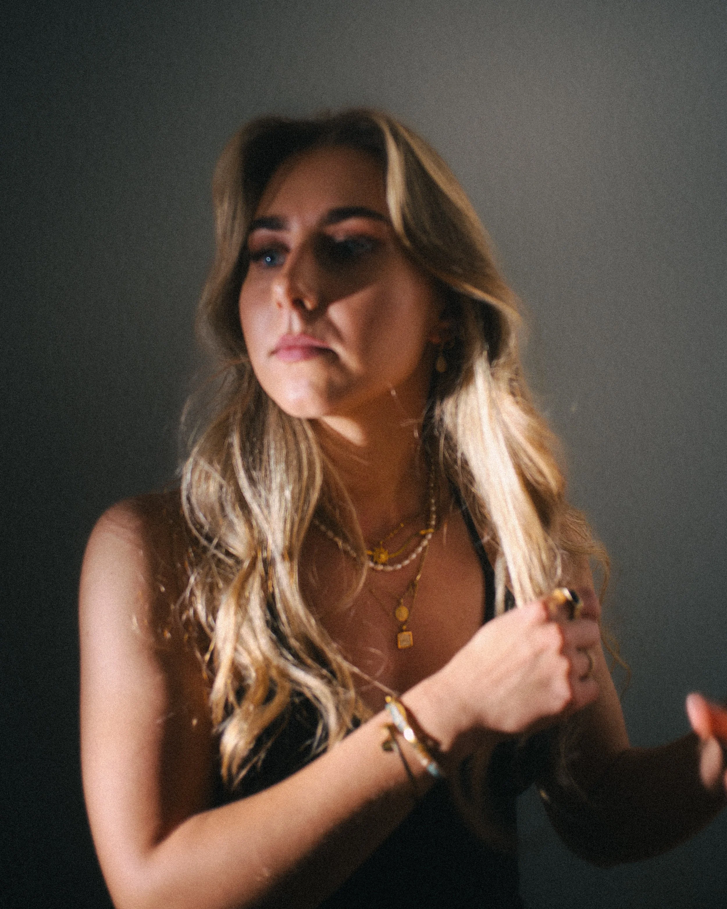 A young woman with wavy blonde hair wearing layered gold necklaces, multiple rings, and bracelets. She is looking slightly to the side with a serious expression, posing against a plain dark background.