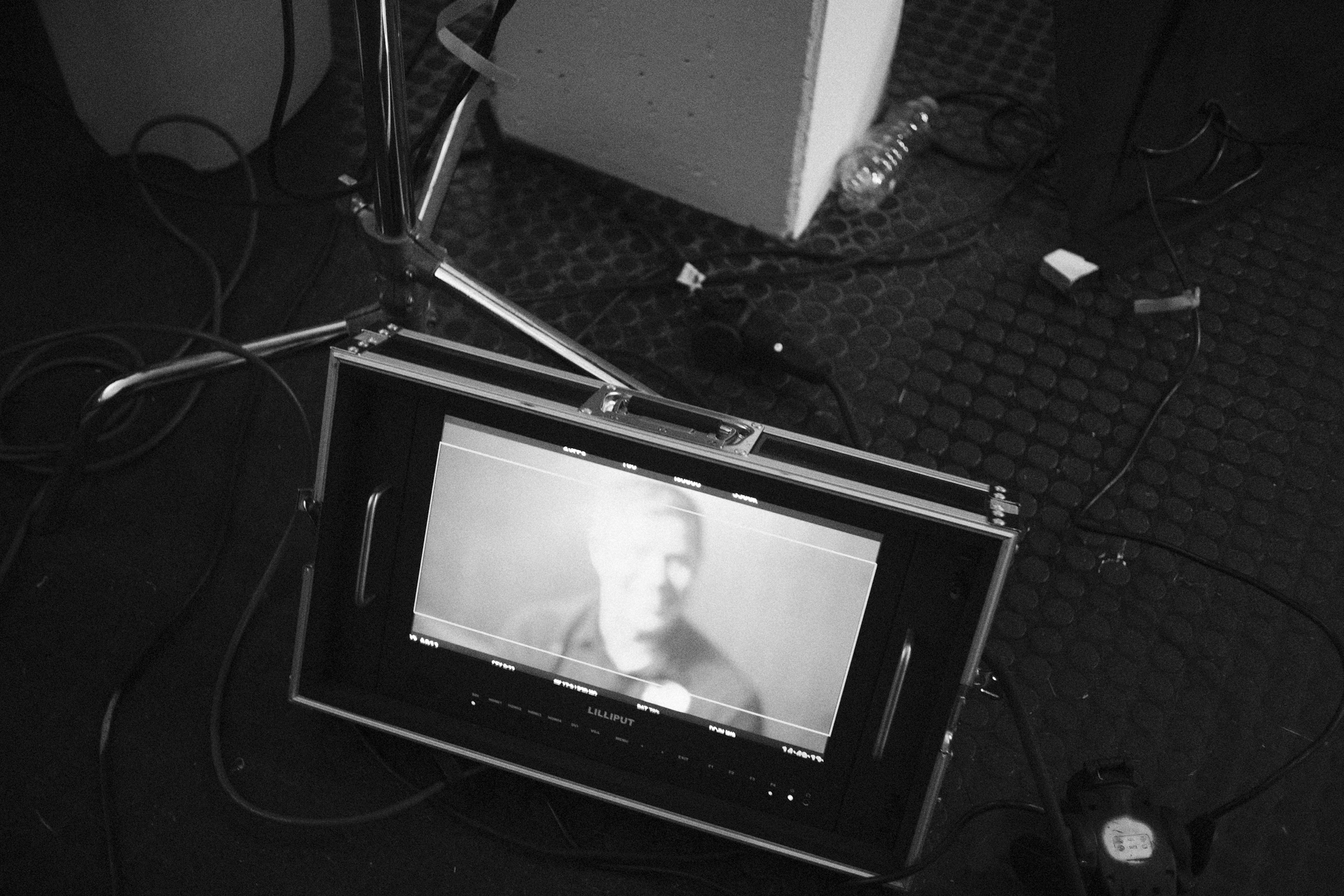 A video monitor displaying a black-and-white video scene with a person's face. The monitor is on a floor with cables around it. Nearby, there is a water bottle, a white box, and some electronic equipment.