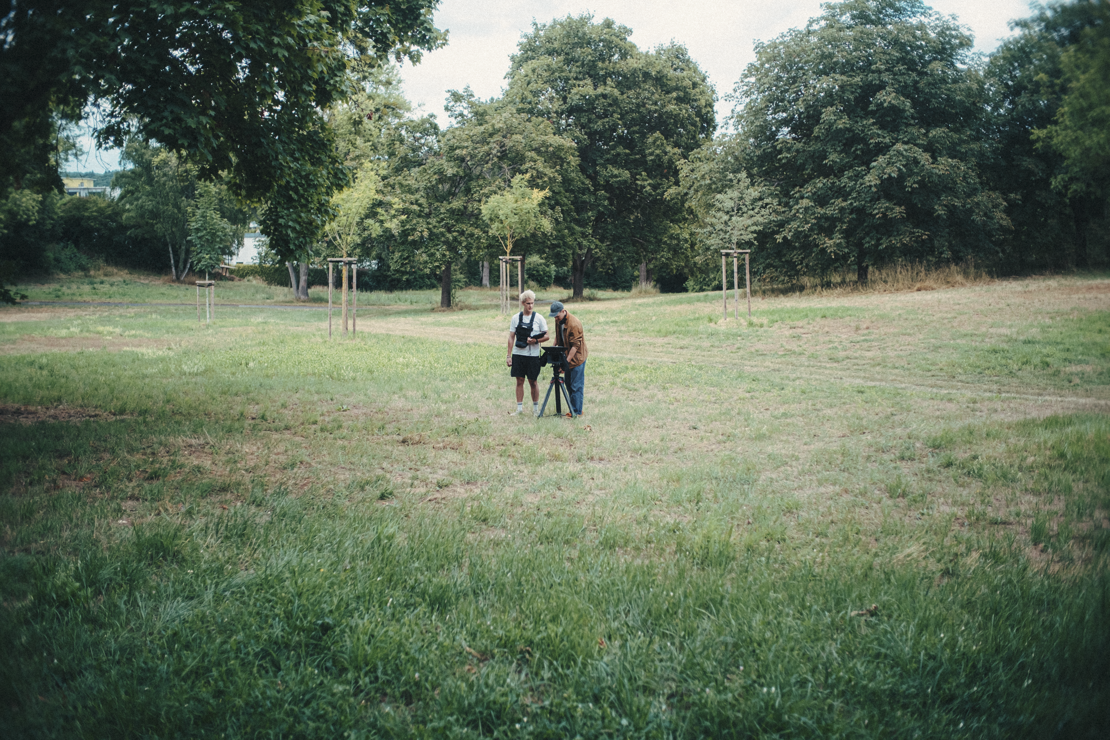 Two people stand in a grassy park, observing a camera on a tripod, surrounded by trees.