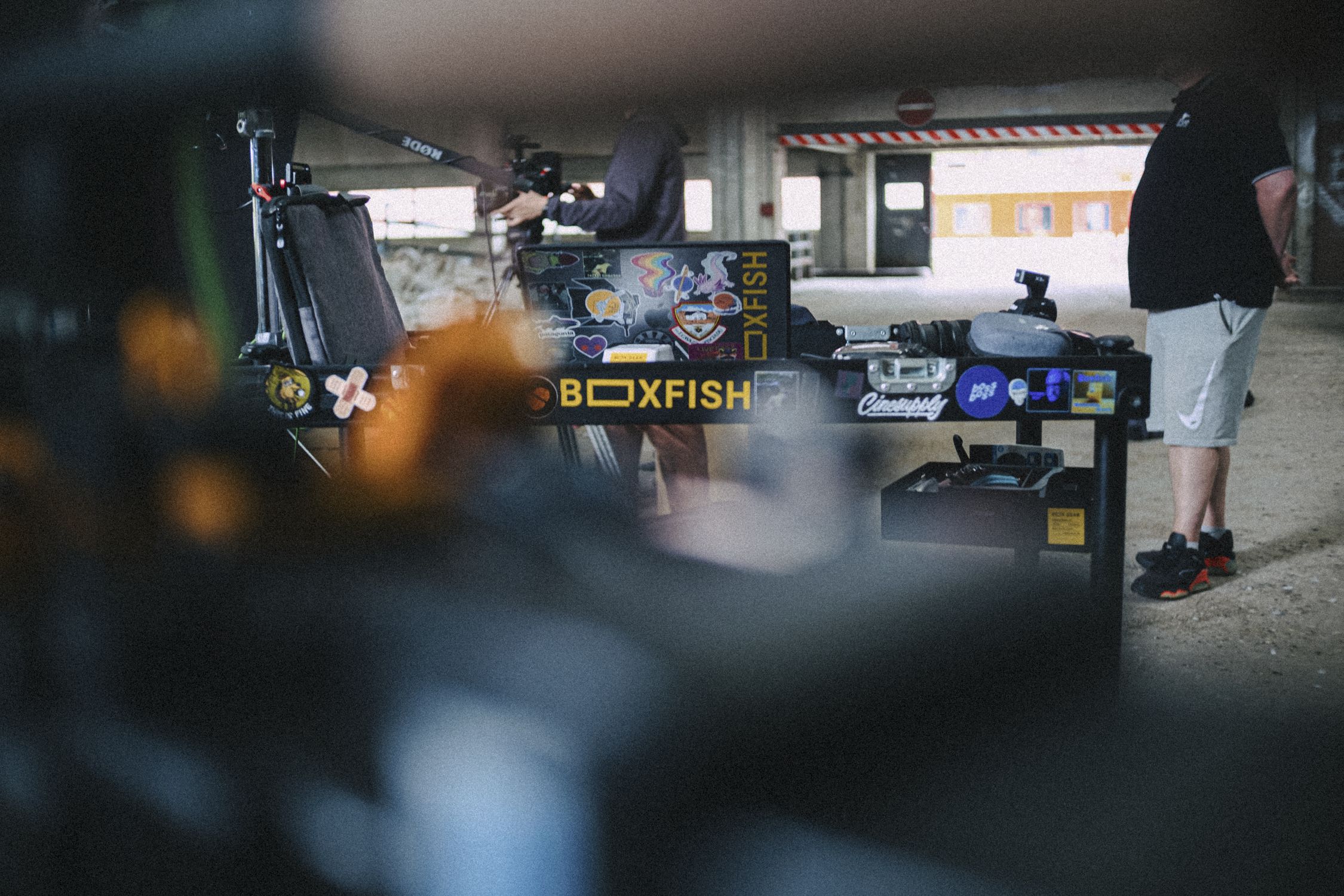 People working on video equipment and cameras under a bridge or parking structure, with various branded stickers and logos on equipment, including 'BOXFISH'.