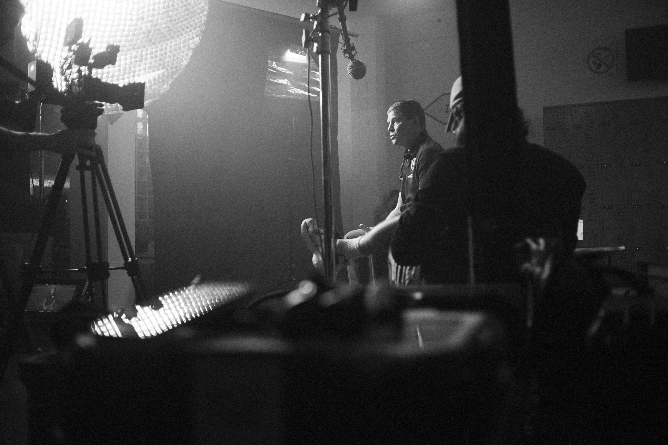 People in a dimly lit room, with a large circular light, filming crew, and a person sitting in front of a backdrop, possibly during an interview or recording session.