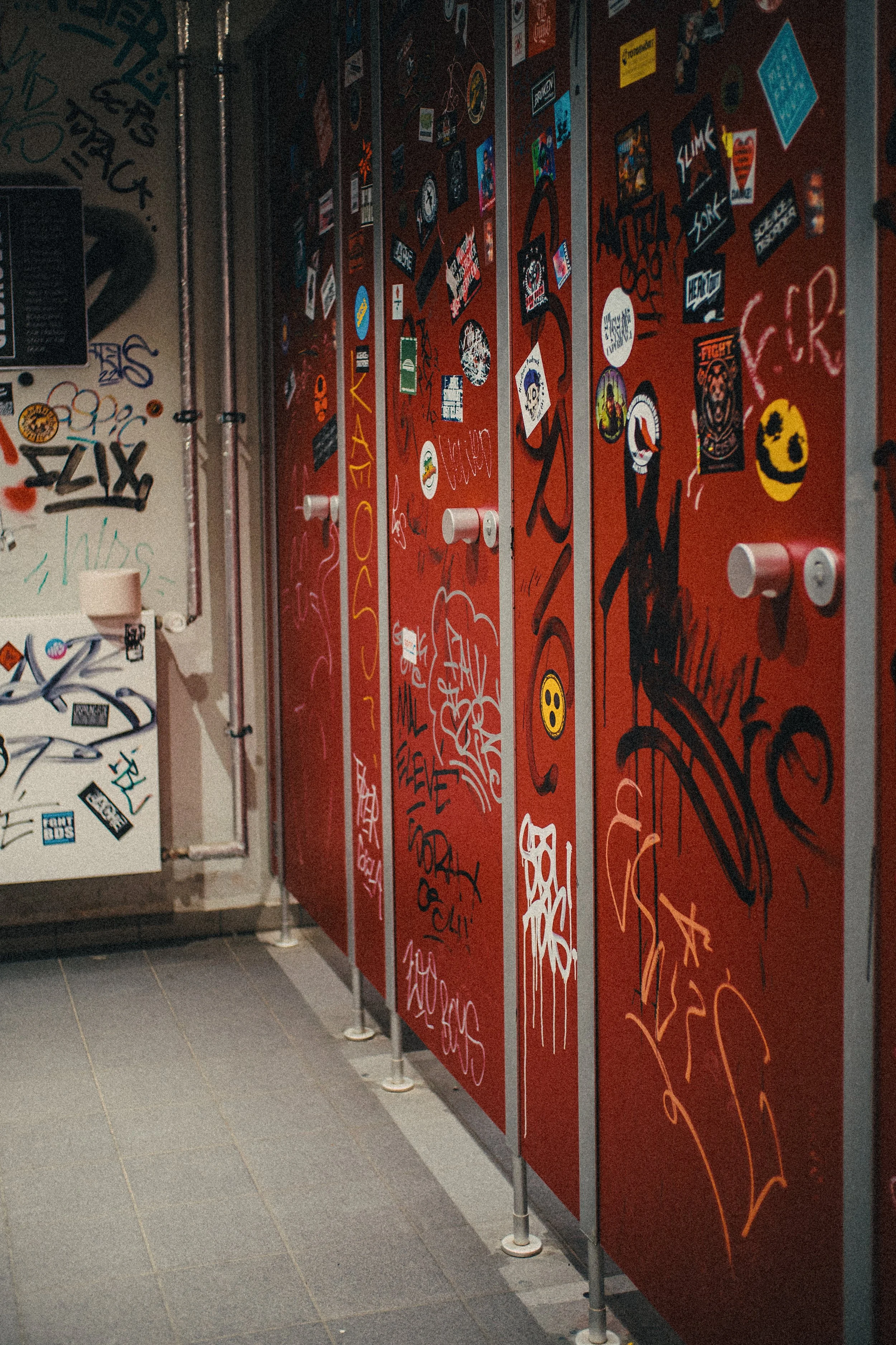 Red locker doors covered in various stickers, graffiti, and graffiti tags in a bathroom or locker room setting.