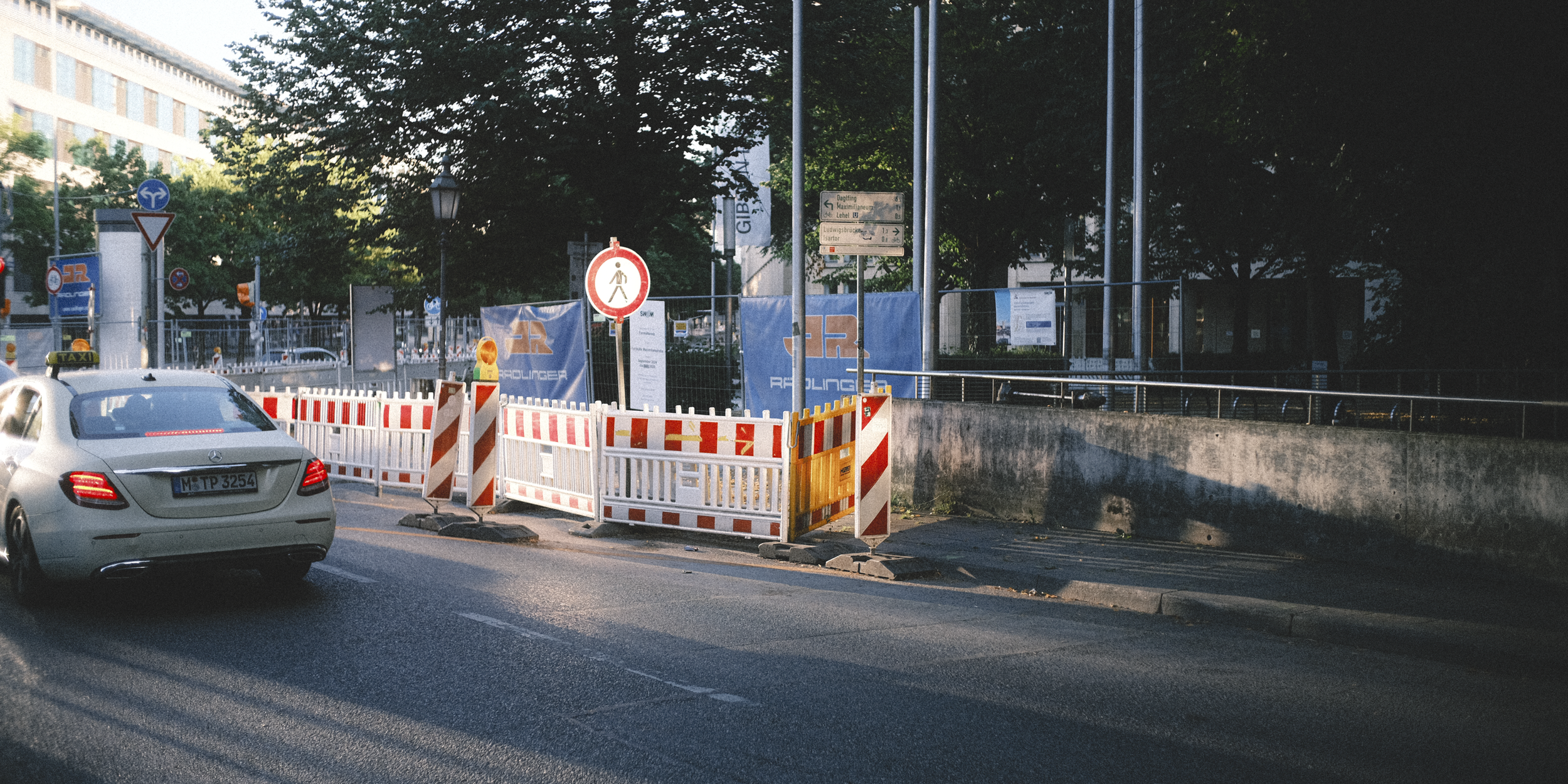 Street view showing a white Mercedes-Benz car with taxi sign, and a sidewalk section cordoned off with orange and white barrier fences, traffic cones, and a no pedestrian sign, with trees and city buildings in the background.