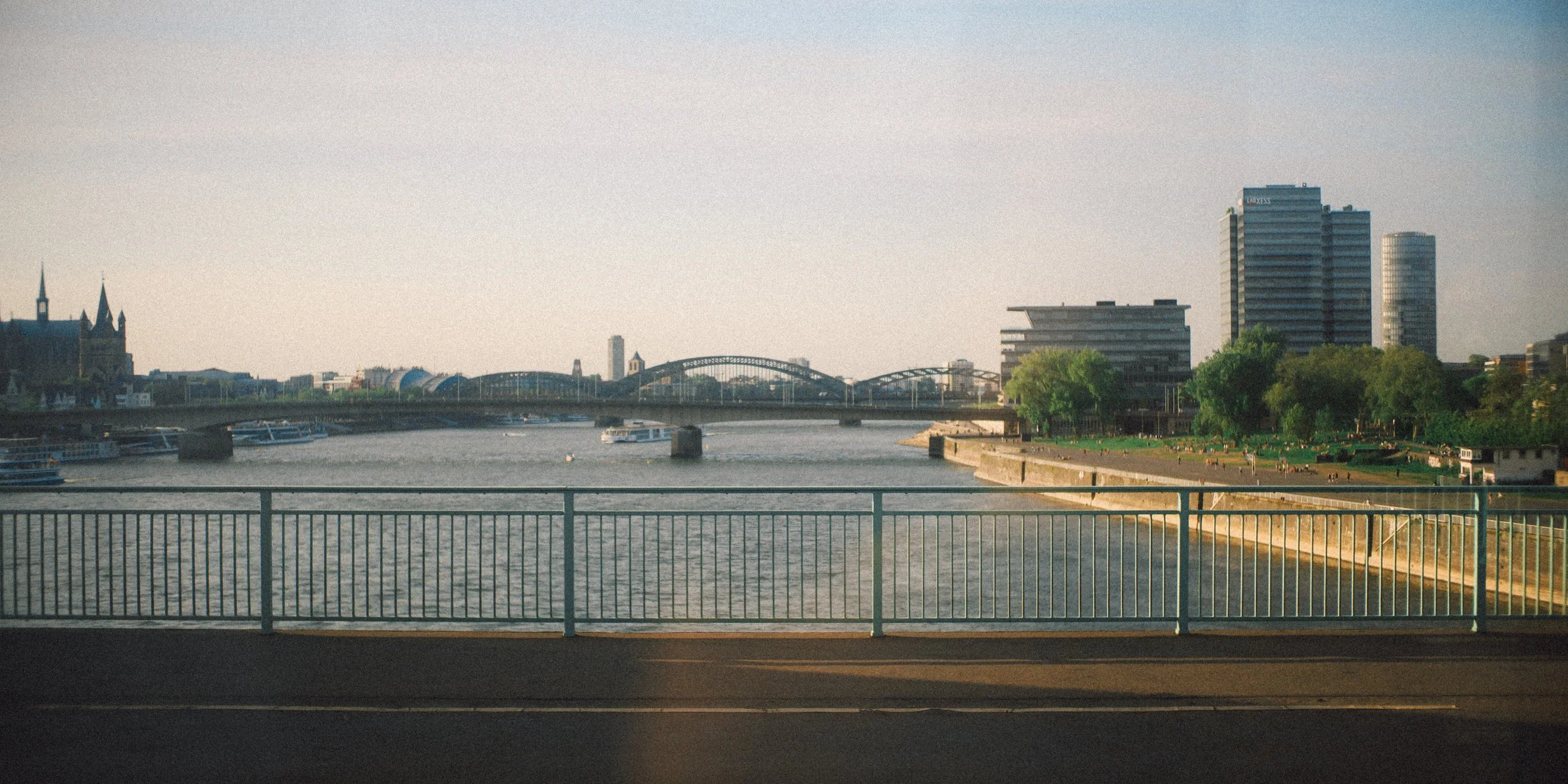 A cityscape view of a river, boats, a bridge, and modern buildings with green trees along the riverbank, taken during daylight.