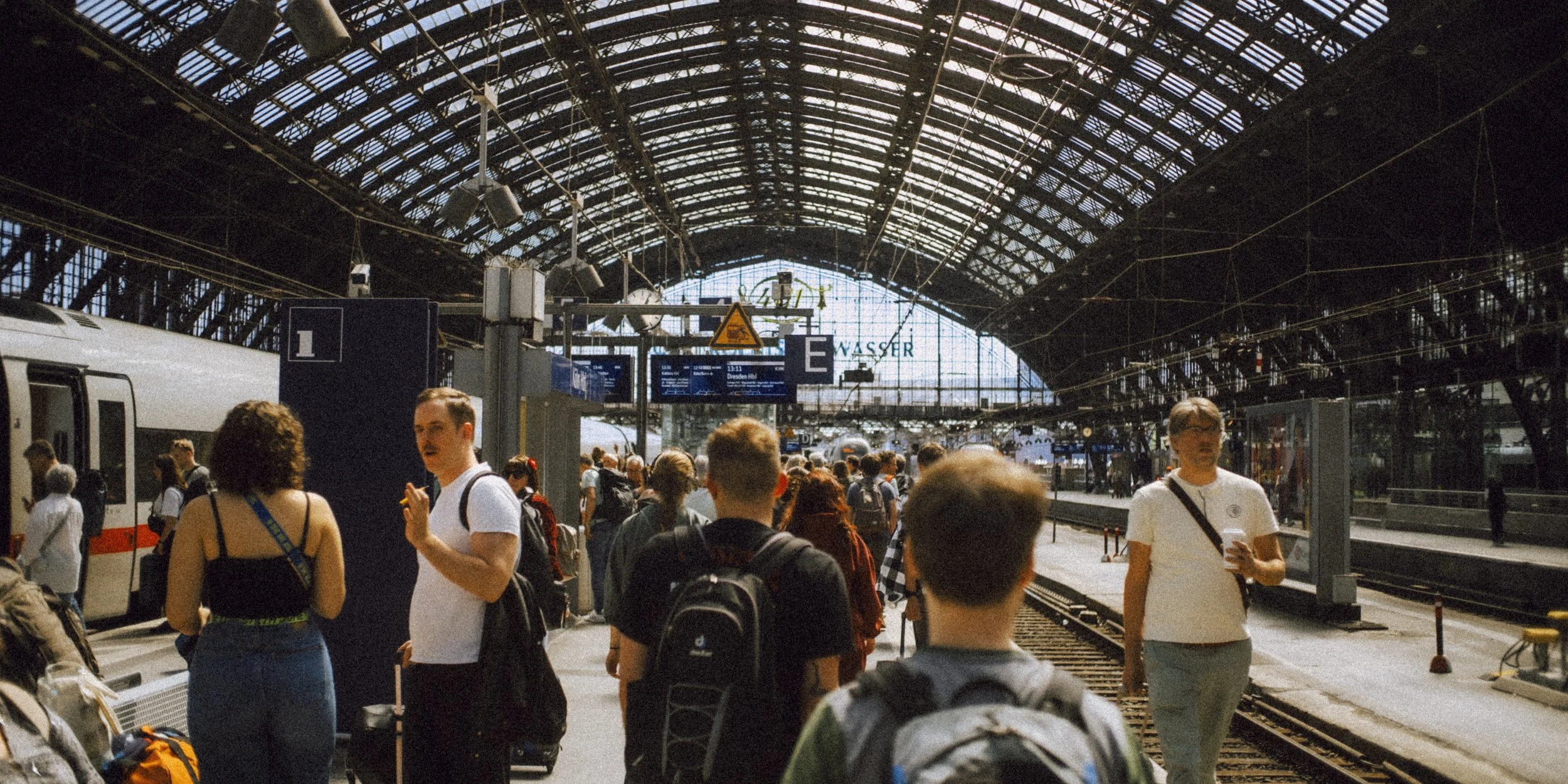Crowd of people at a train station with a high arched glass ceiling, some holding luggage and others using phones, near train tracks and platforms.