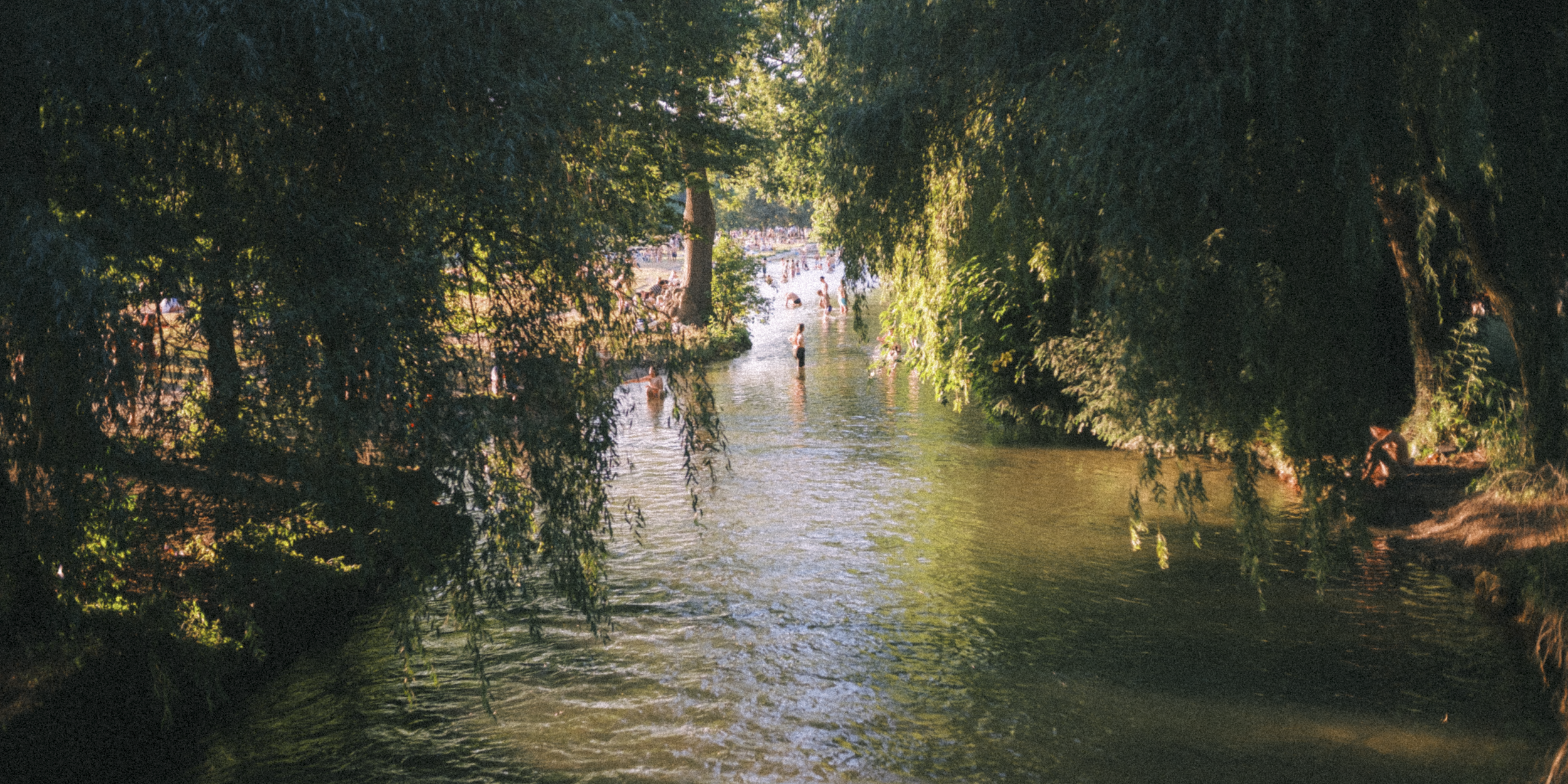 People swimming and wading in a river surrounded by trees and greenery, with sunlight filtering through the leaves.