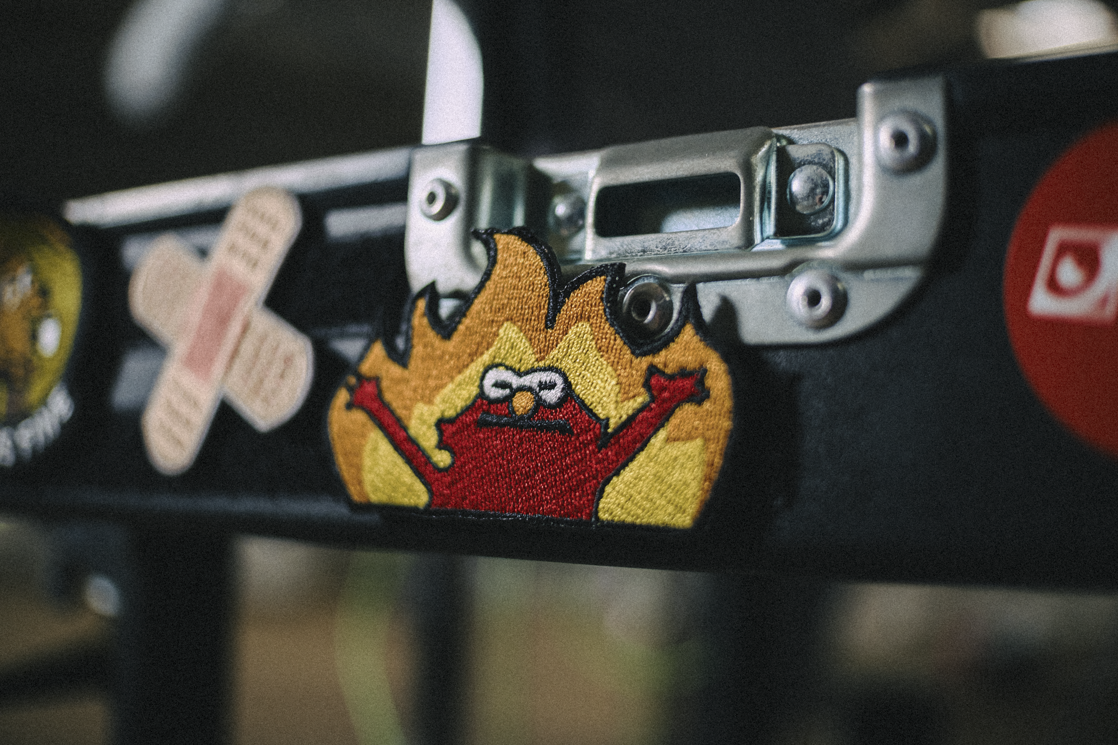 Close-up of a black metal surface with a colorful embroidered patch of a red character with glasses and arms raised, along with other patches including a bandage design.
