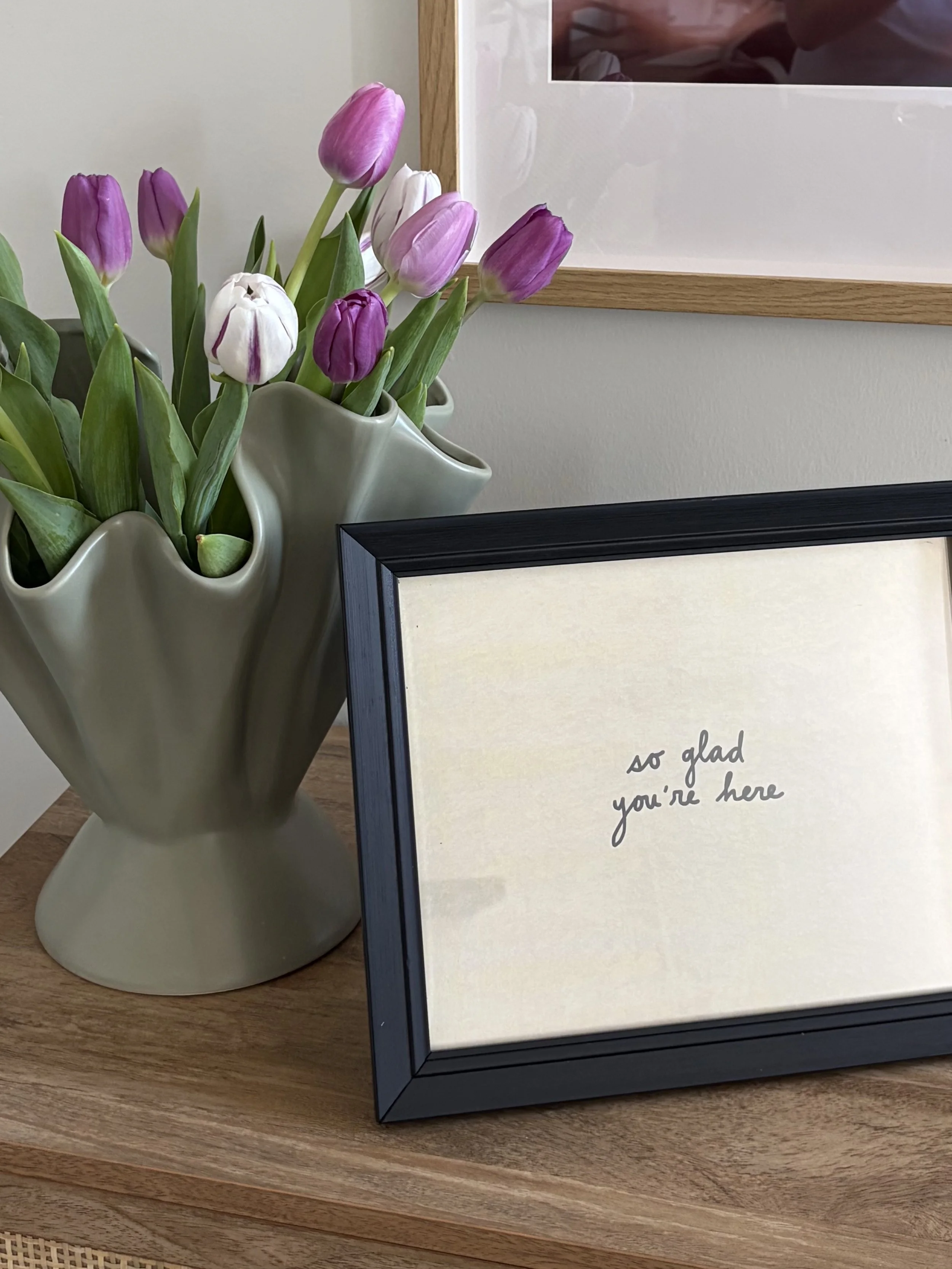 A white ceramic vase with a wavy design holding a bouquet of purple and white tulips, placed next to a framed quote that says, 'so glad you're here,' on a wooden surface.