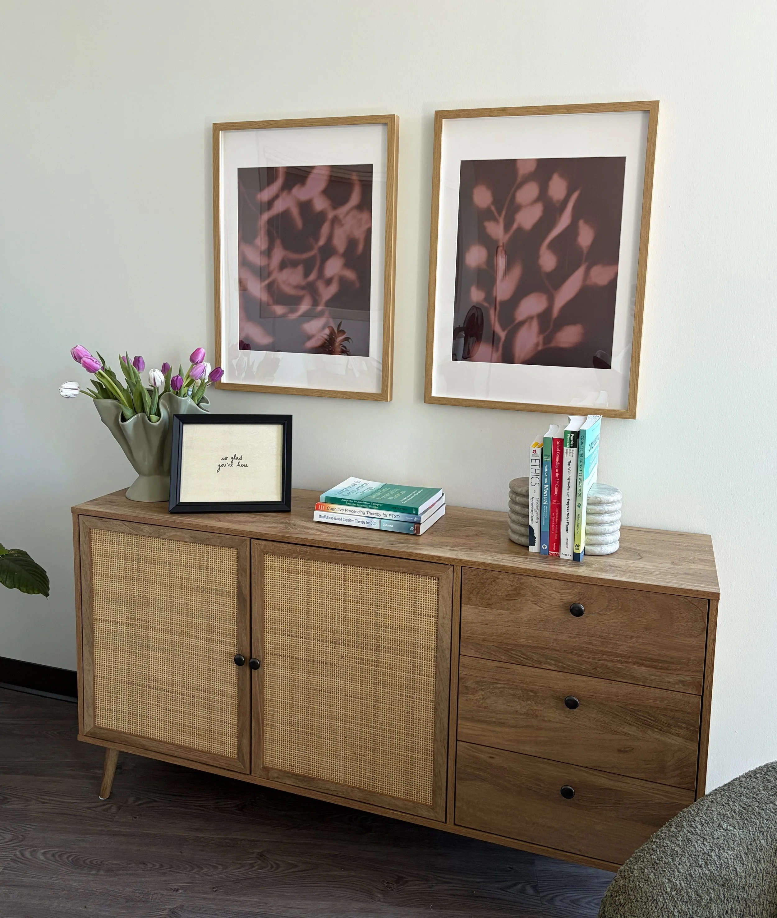 A wooden sideboard with two cabinet doors featuring woven rattan panels and four drawers on the right side. On top of the sideboard are a potted plant with purple and white tulips, a stack of books, and a framed quote saying "so glad you're here." Behind the sideboard, the wall displays two framed abstract pictures with pinkish-brown organic shapes. The floor is dark wood, and part of a gray upholstered chair or bench is visible in the lower right corner.