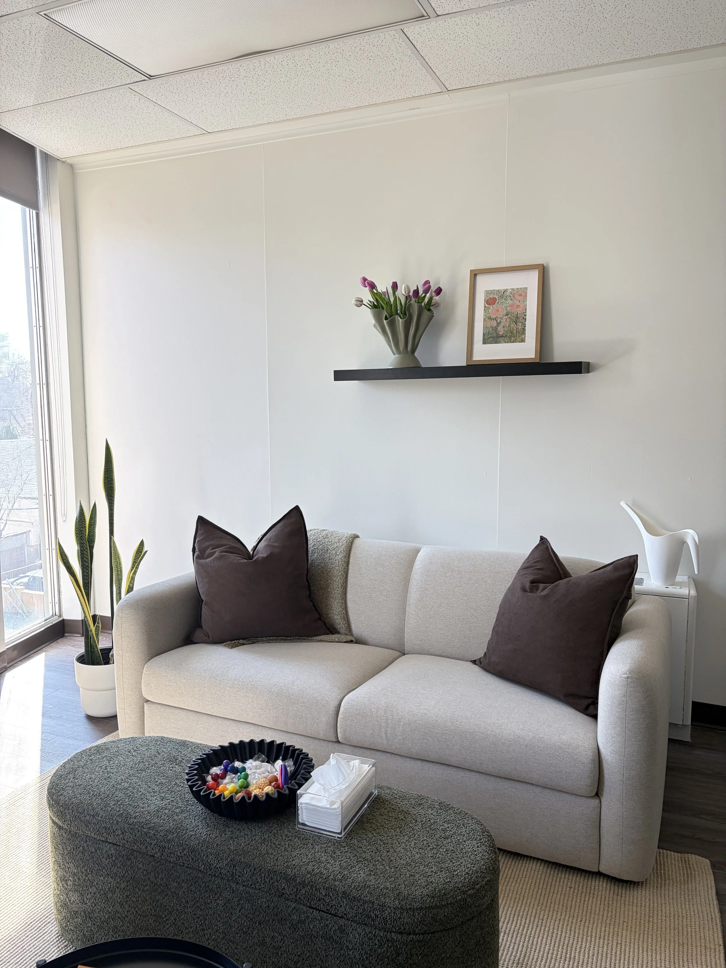 Living room with a beige sofa and dark brown pillows, a black round coffee table with colorful candies and tissues, a tall potted plant near the window, a wall shelf with a vase of pink and purple tulips and a framed floral picture, a white side table with a white pitcher, and large window with blinds.