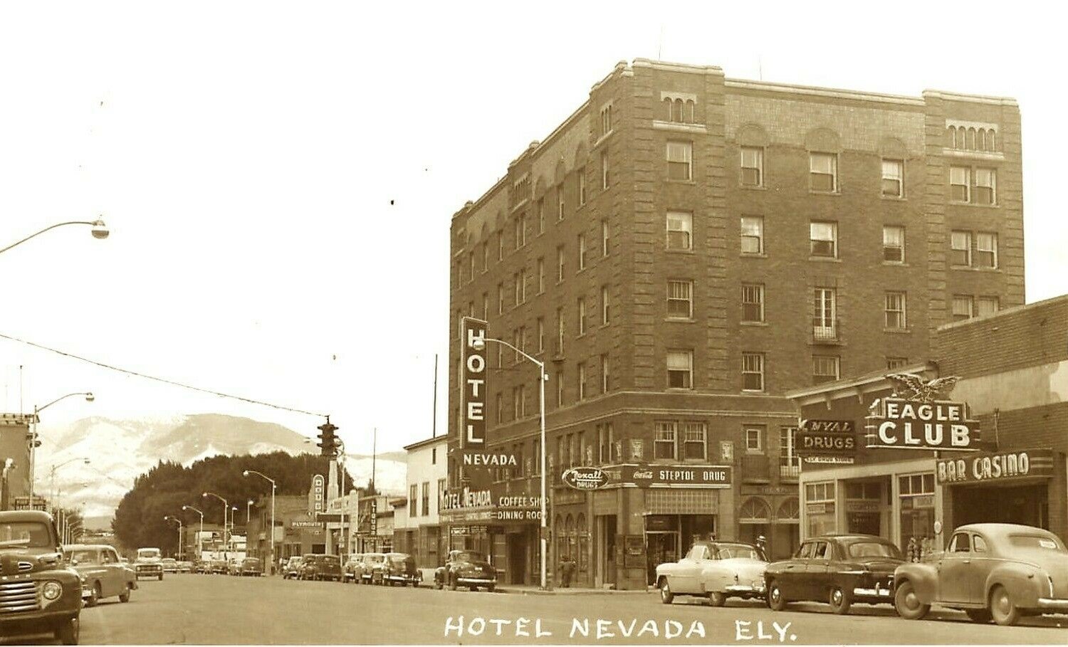 Vintage black-and-white photograph of Hotel Nevada, a tall brick building, on a busy street with parked cars, multiple signs including a neon 'Eagle Club', a streetlamp, and hills or mountains in the background.