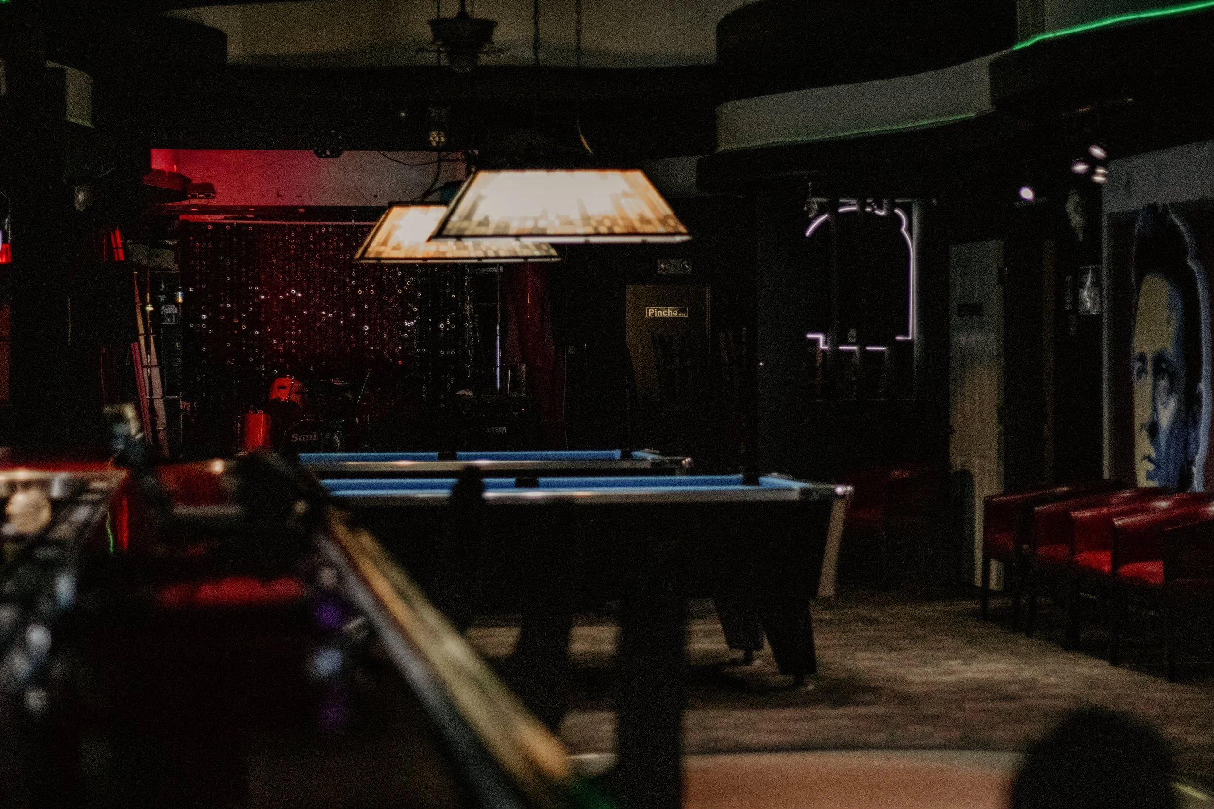 Dimly lit bar or music venue with tables, hanging pendant lights, and a decorated backdrop backdrop, possibly for live performances.