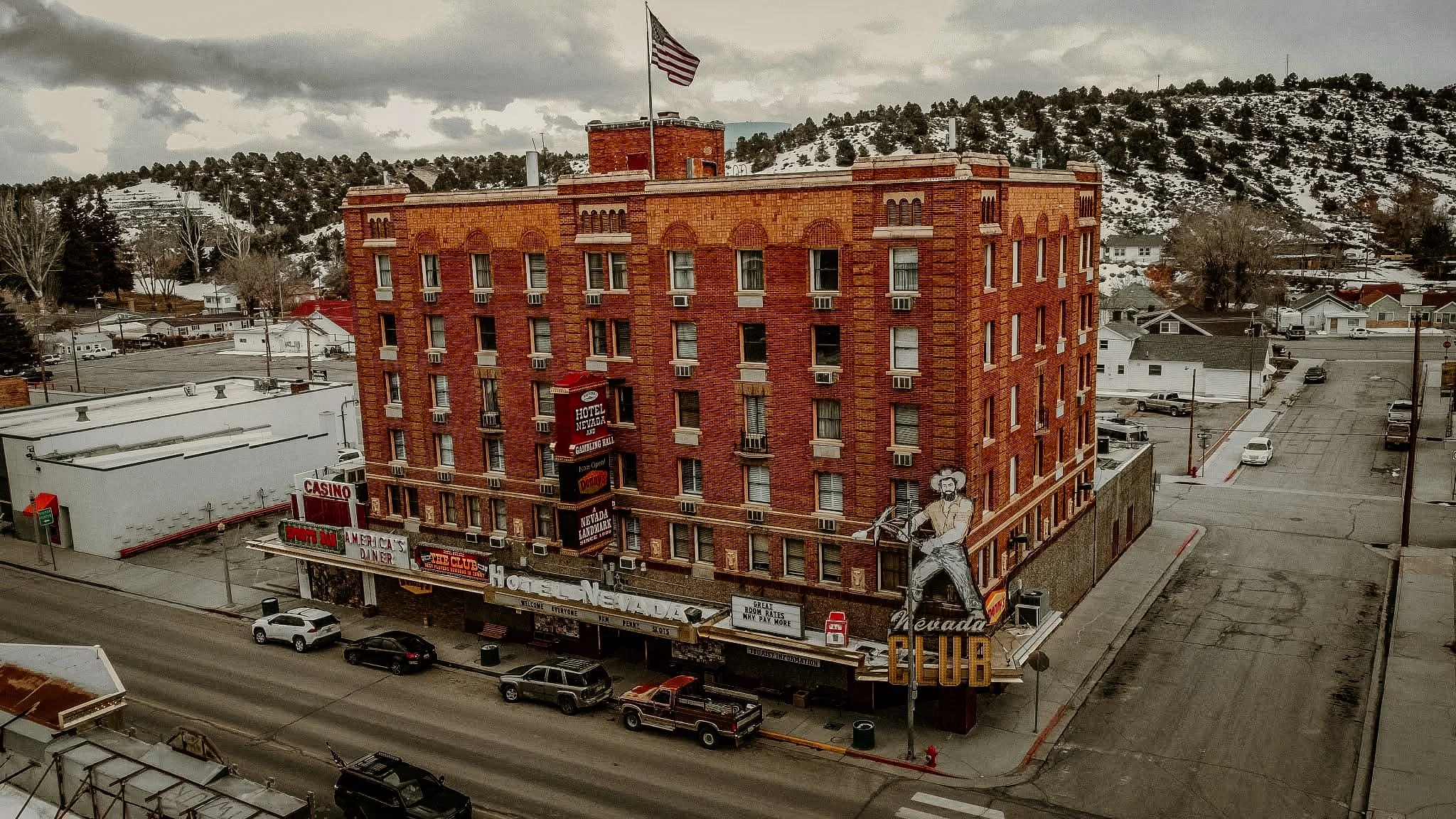 A multi-story red brick hotel building with an American flag on top, surrounded by snow-covered hills and streets with parked cars and small commercial establishments in a small town setting.