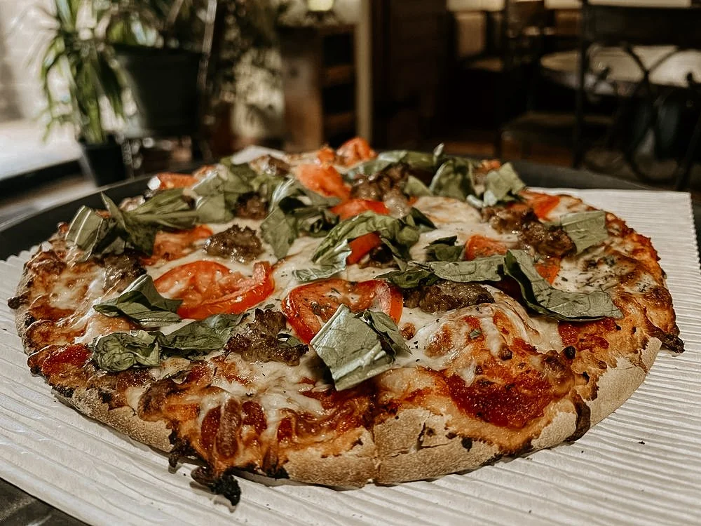 A freshly baked pizza topped with cheese, basil, tomato slices, and cooked sausage slices on a wooden table.