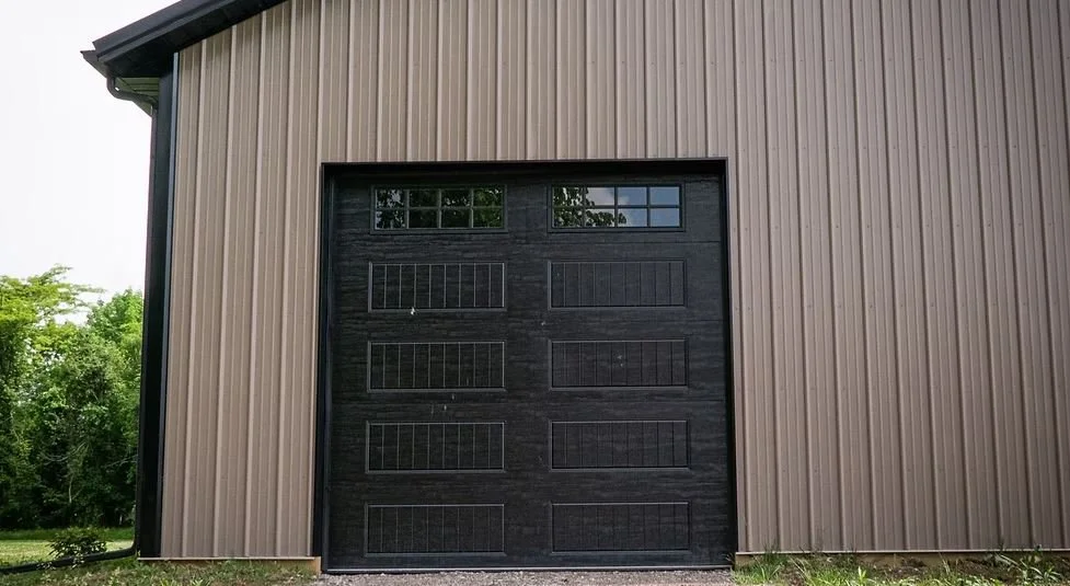 A large black garage door set against a beige metal building exterior, with small windows at the top and surrounded by green trees and grass.