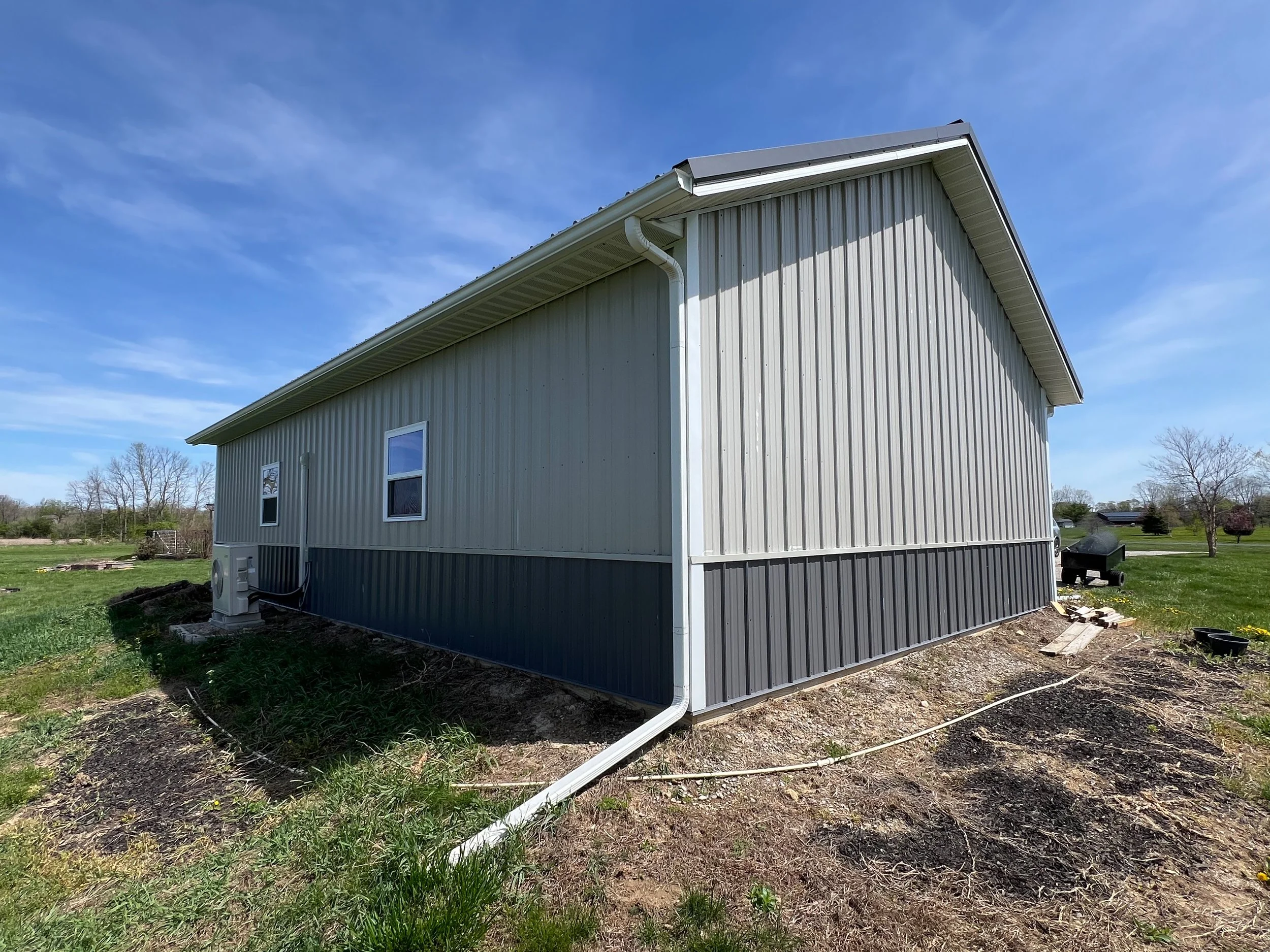 Exterior of a light-colored metal house with a gable roof, showing primarily the side view with a gutter system, a small window, and some electrical equipment near the foundation. The house is situated on a grassy area with some trees and a partly cl