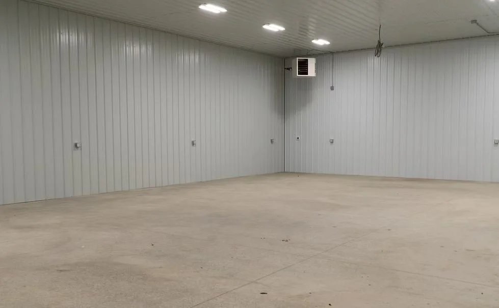 Empty interior of a large room with light gray metal walls and ceiling, a beige concrete floor, fluorescent ceiling lights, and a wall-mounted heater or vent in the corner.