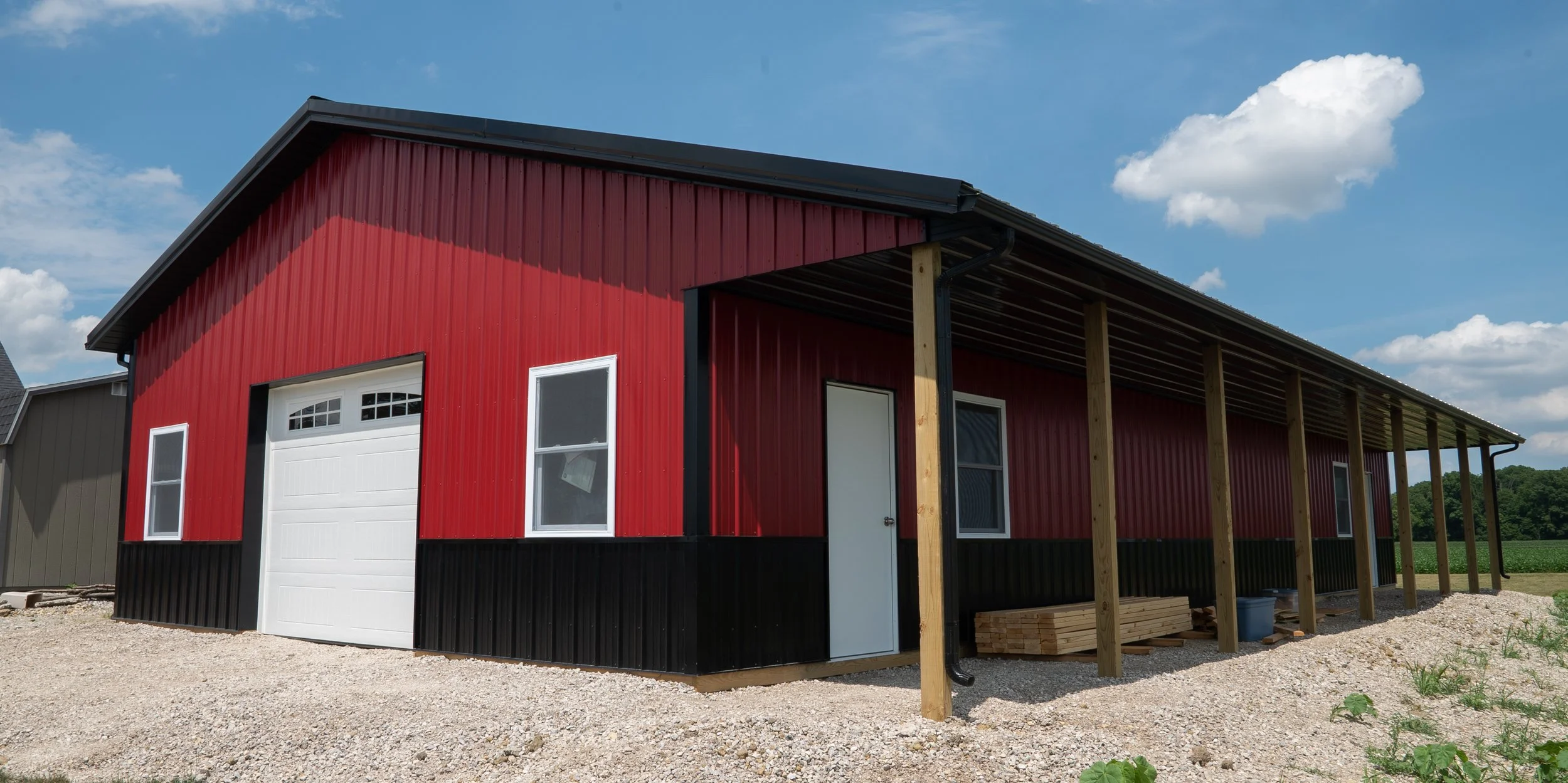 A red metal building under construction with a white garage door, windows, and a side door. The building is elevated on wooden posts, and there is gravel around it with some greenery. The sky is blue with a few clouds.