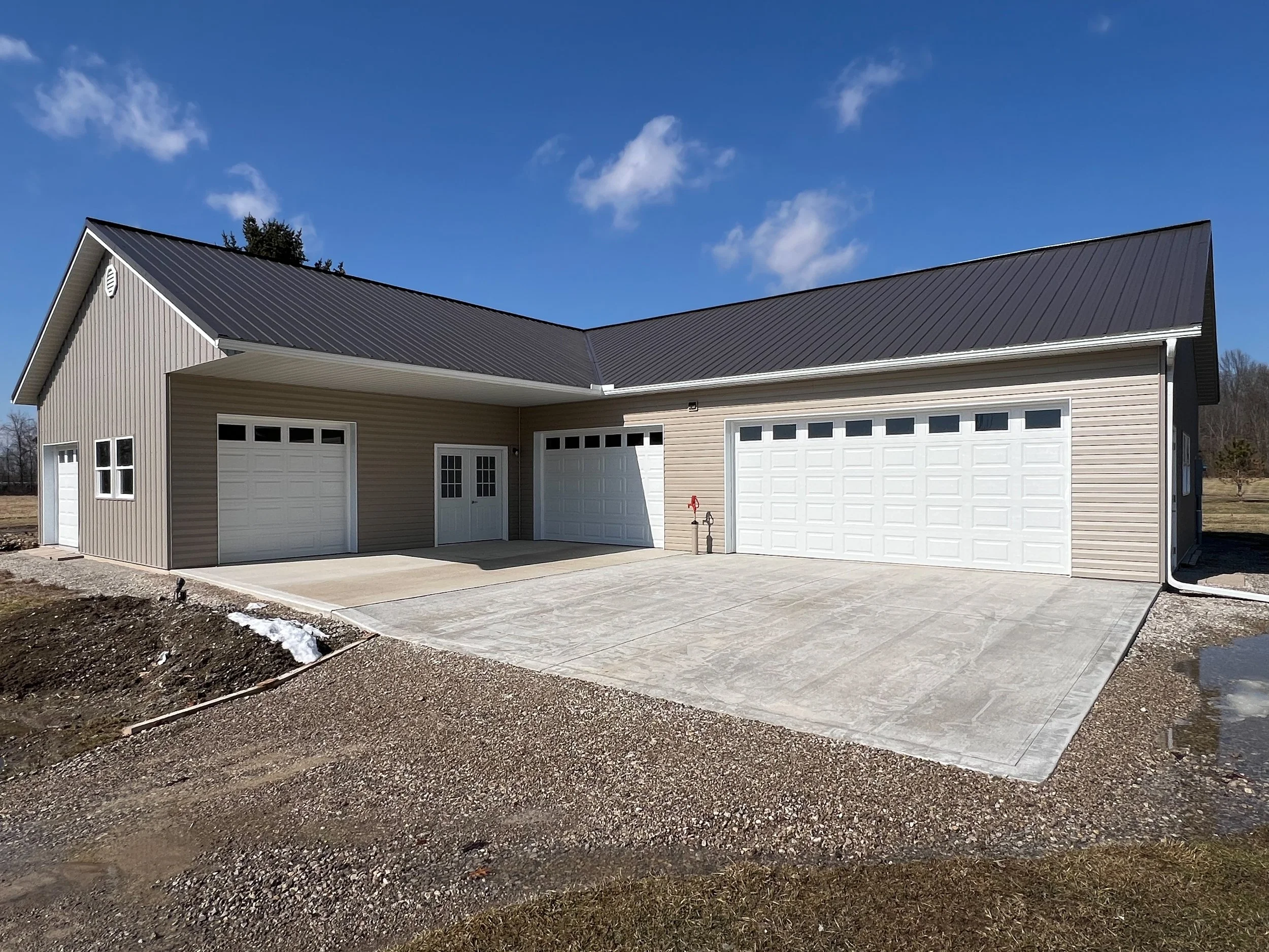 A modern house with a beige exterior and a metal roof, featuring three white garage doors and a concrete driveway, under a clear blue sky.