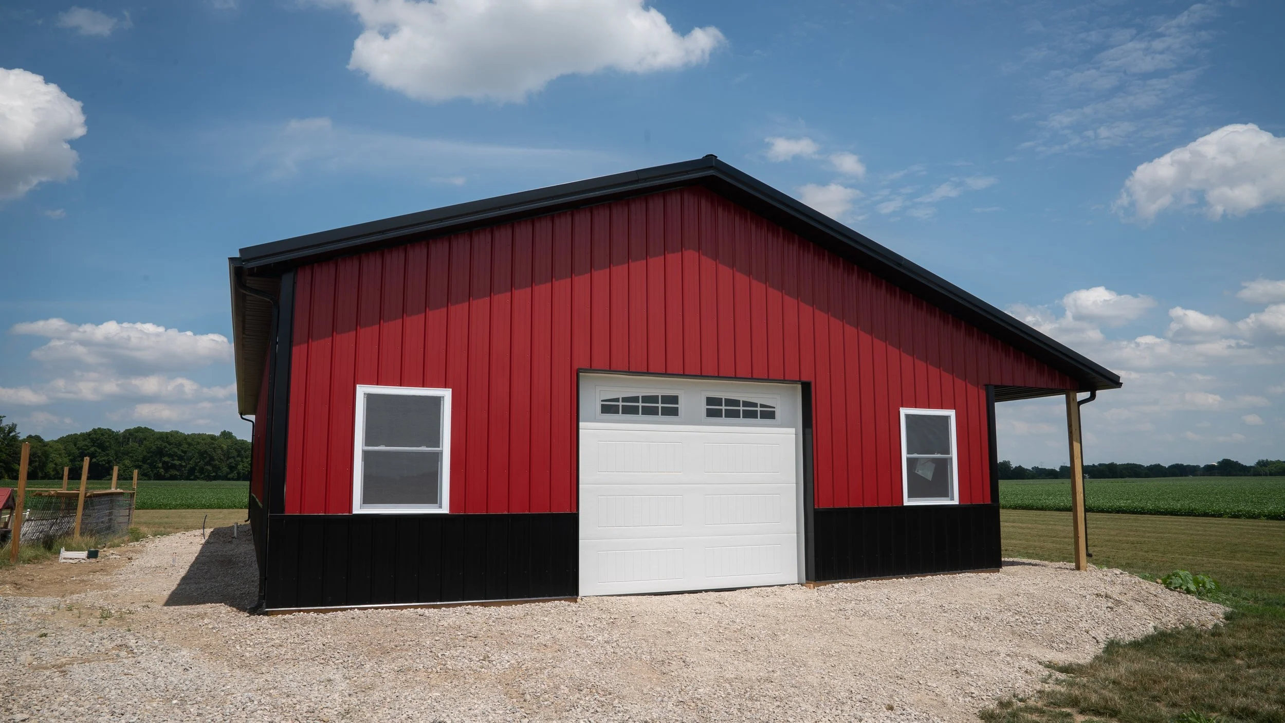 Red barn with white garage door and windows, set on a gravel lot under a partly cloudy sky with green fields in the background.
