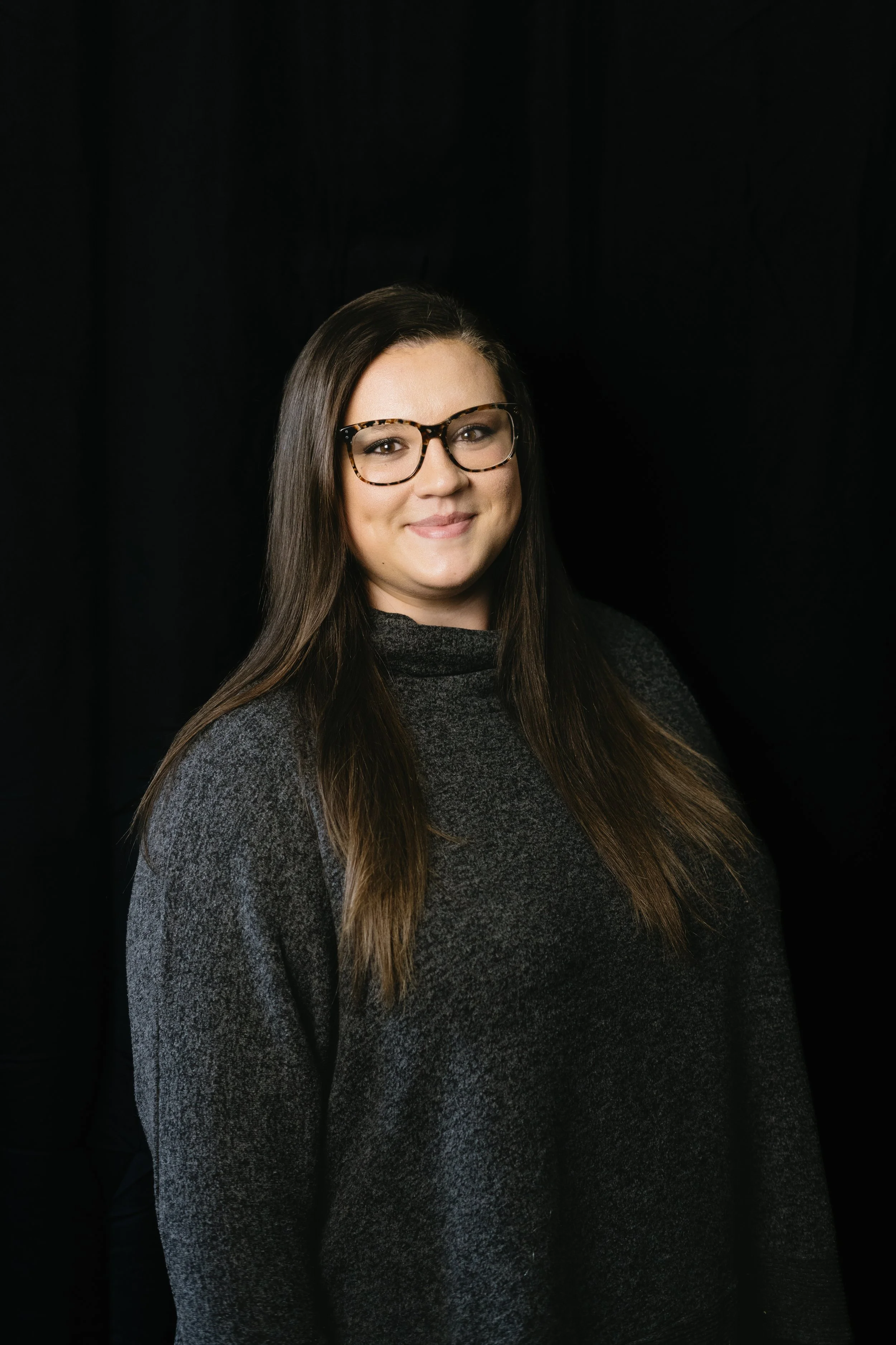 A young woman with long brown hair, wearing glasses with tortoiseshell frames, smiling at the camera, against a black background.
