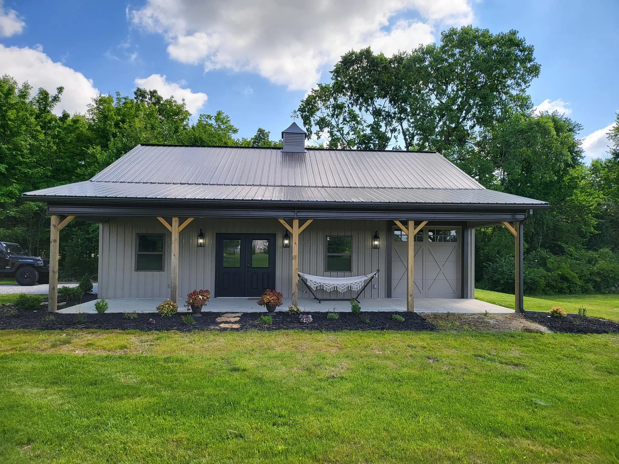 Front view of a small building with a metal roof, wooden support beams, and a decorated hammock in front, surrounded by a well-maintained garden with flowers, green grass, and trees, under a partly cloudy sky.