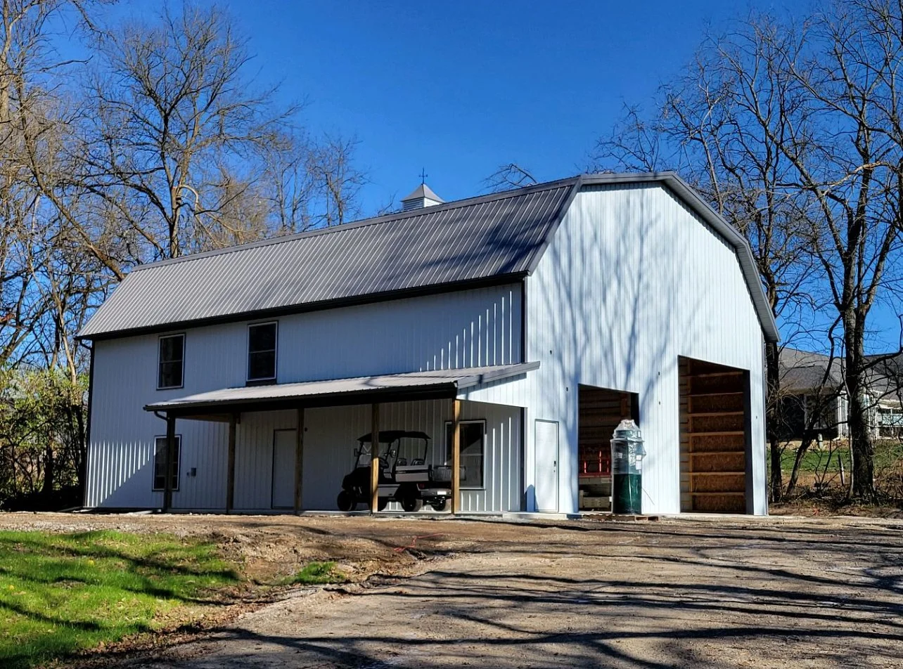 A white barn with a gray metal roof, a covered porch, and a golf cart inside, set against a backdrop of leafless trees and a clear blue sky.
