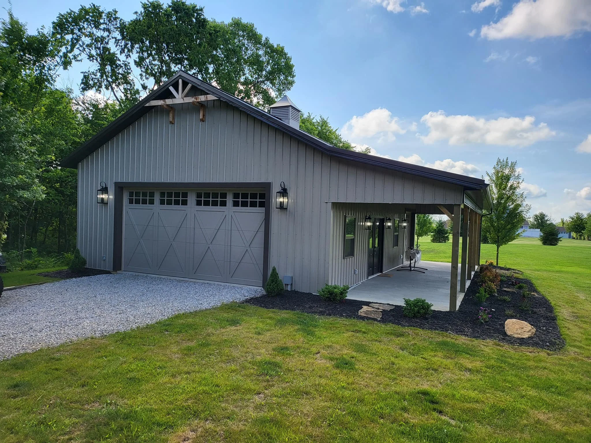 A modern barn with a white paneled garage door and outdoor lighting, surrounded by a well-maintained lawn, trees, and a clear blue sky with scattered clouds.